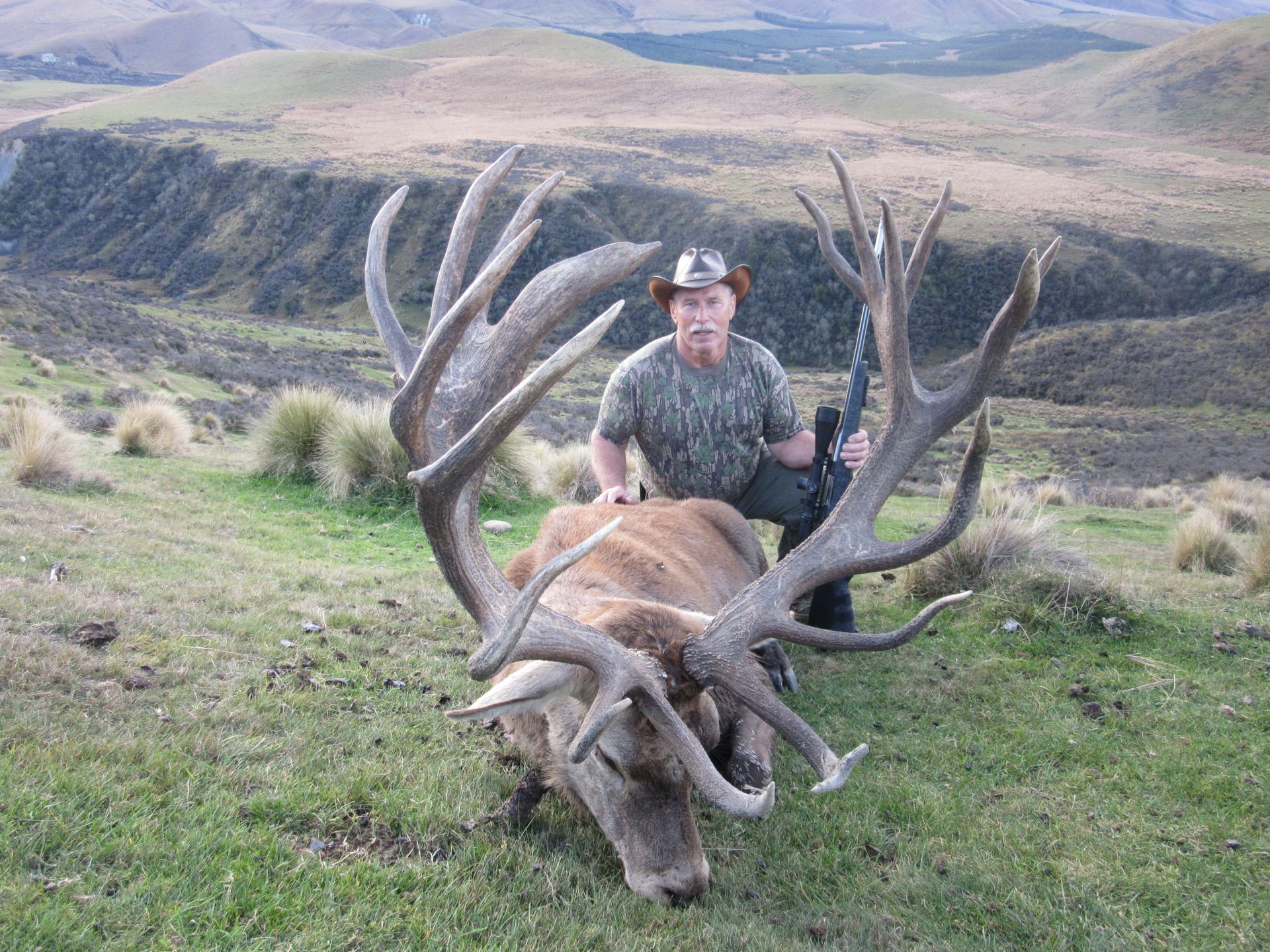 Hunter kneels next to a large dead stag with massive antlers in a grassy hillside setting. He is holding a rifle.