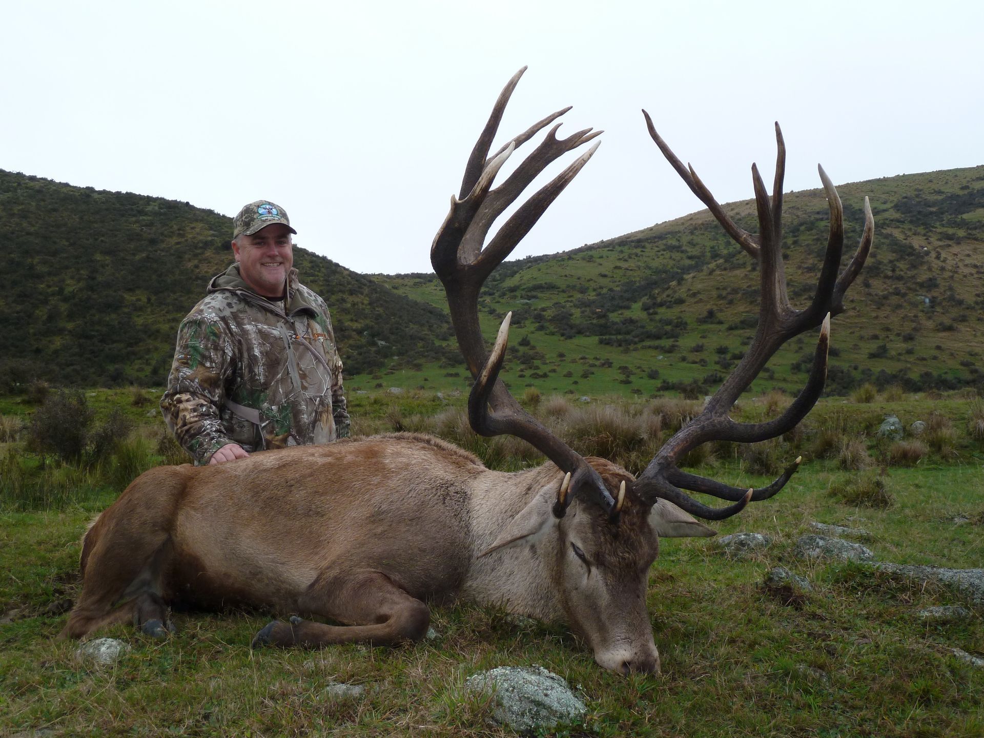 Man in camouflage stands beside a large red deer with impressive antlers, lying on grass in a hilly landscape.