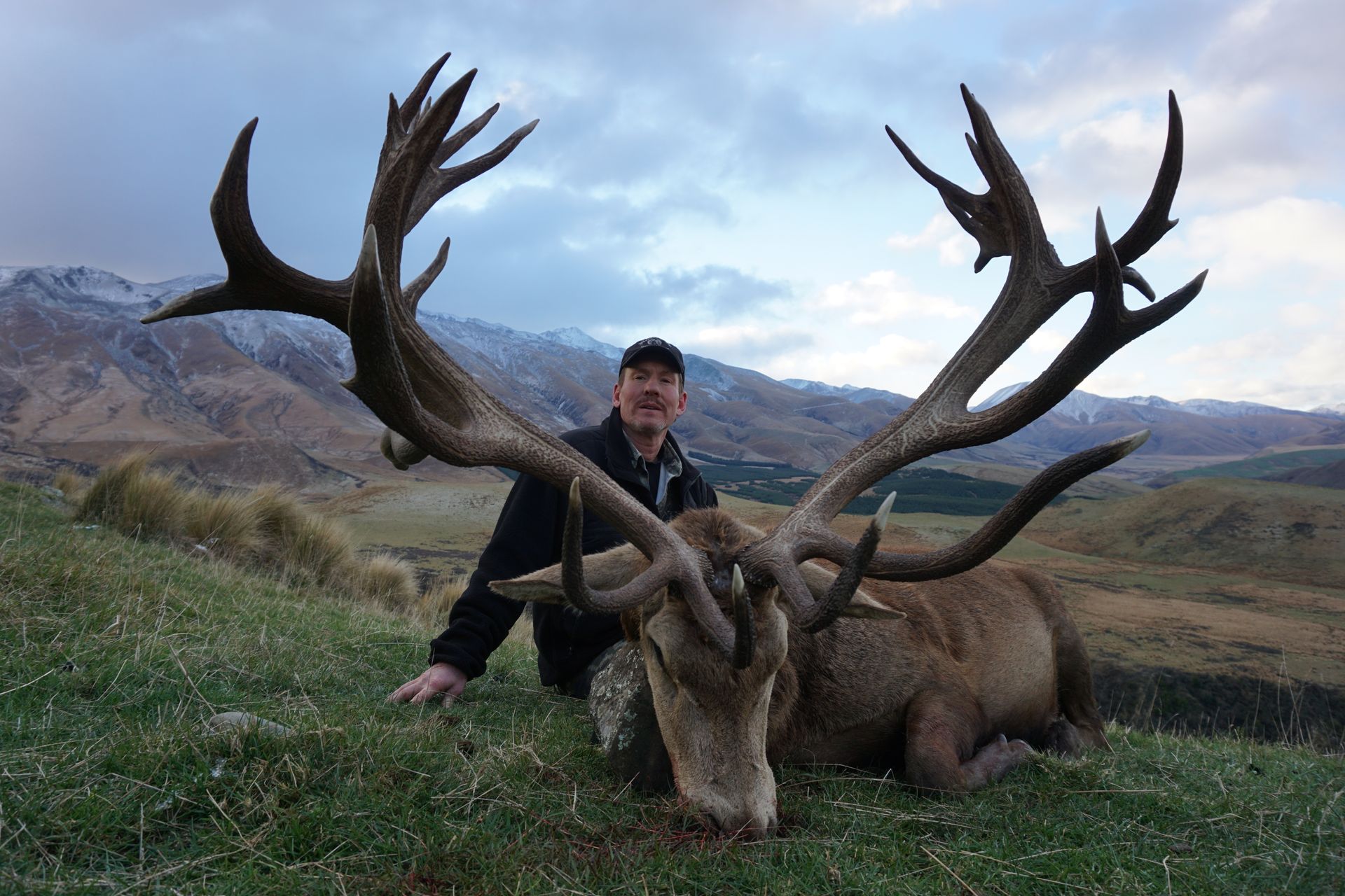 Man kneeling next to a large, dead red deer with impressive antlers, in a grassy mountain landscape under a cloudy sky.