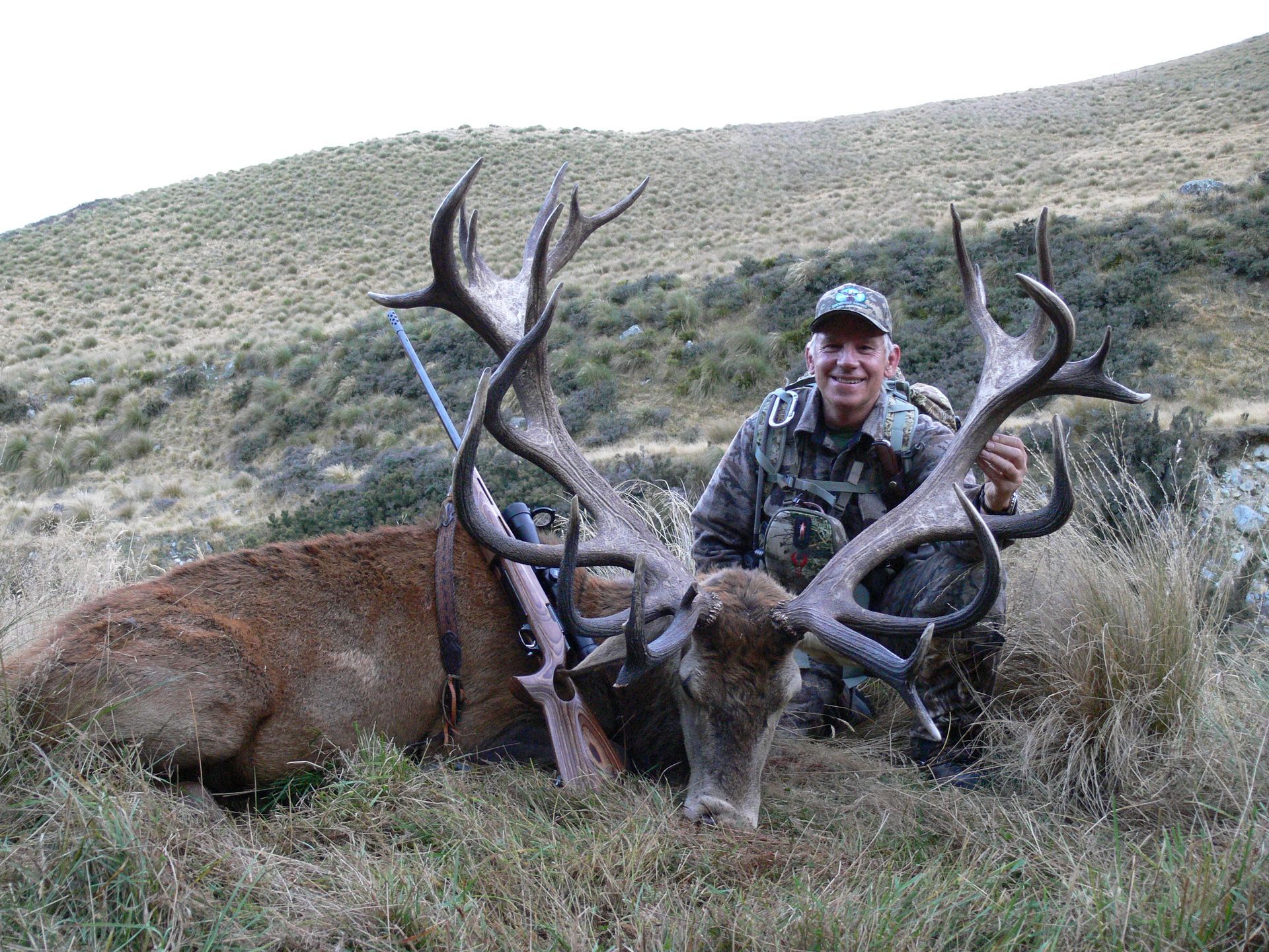Man in camo kneels beside a large dead red deer stag in a grassy, mountainous setting. The man is smiling.