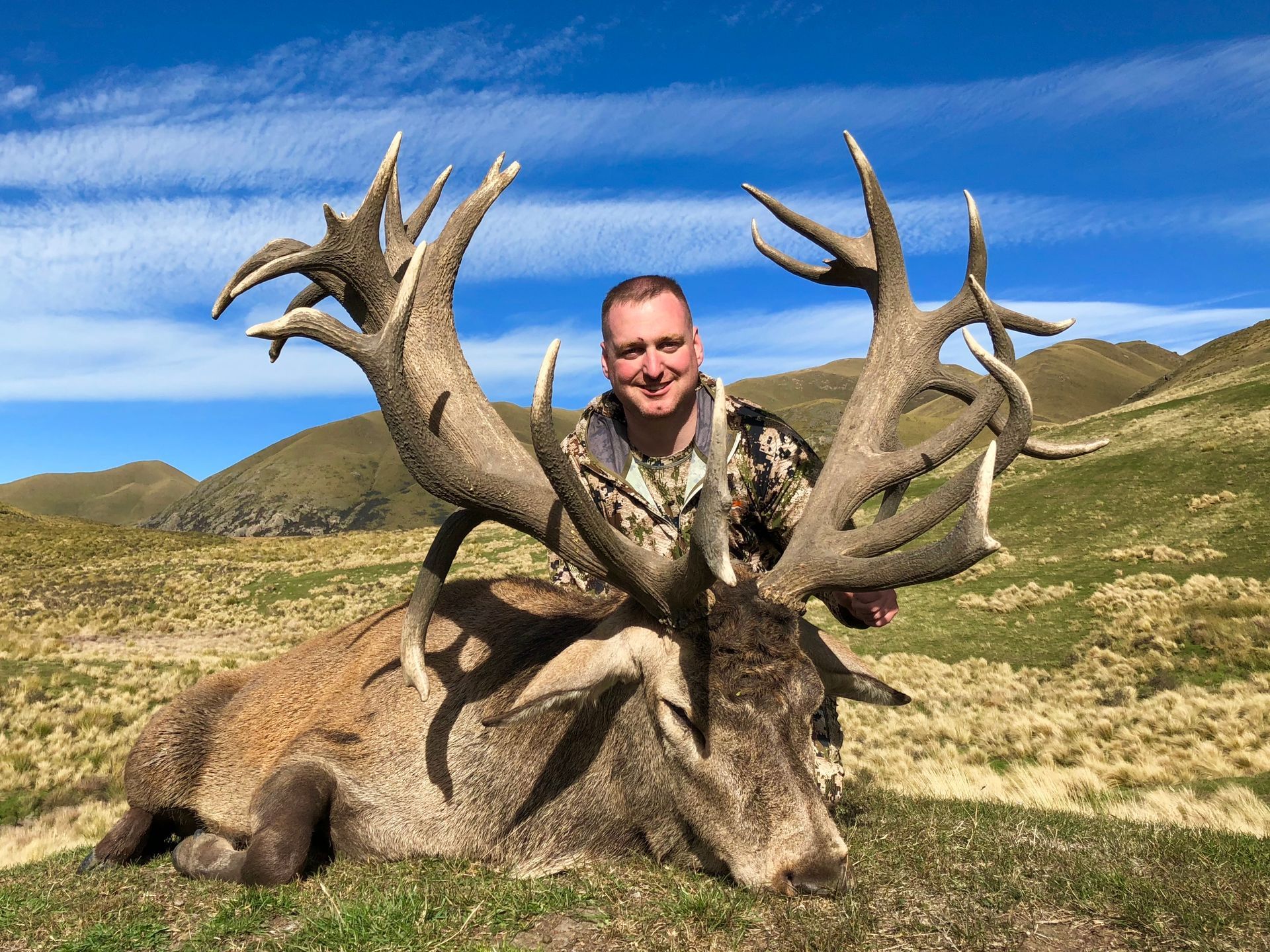 Man smiles next to a large red deer with enormous antlers in a mountainous, grassy setting. He wears camouflage.