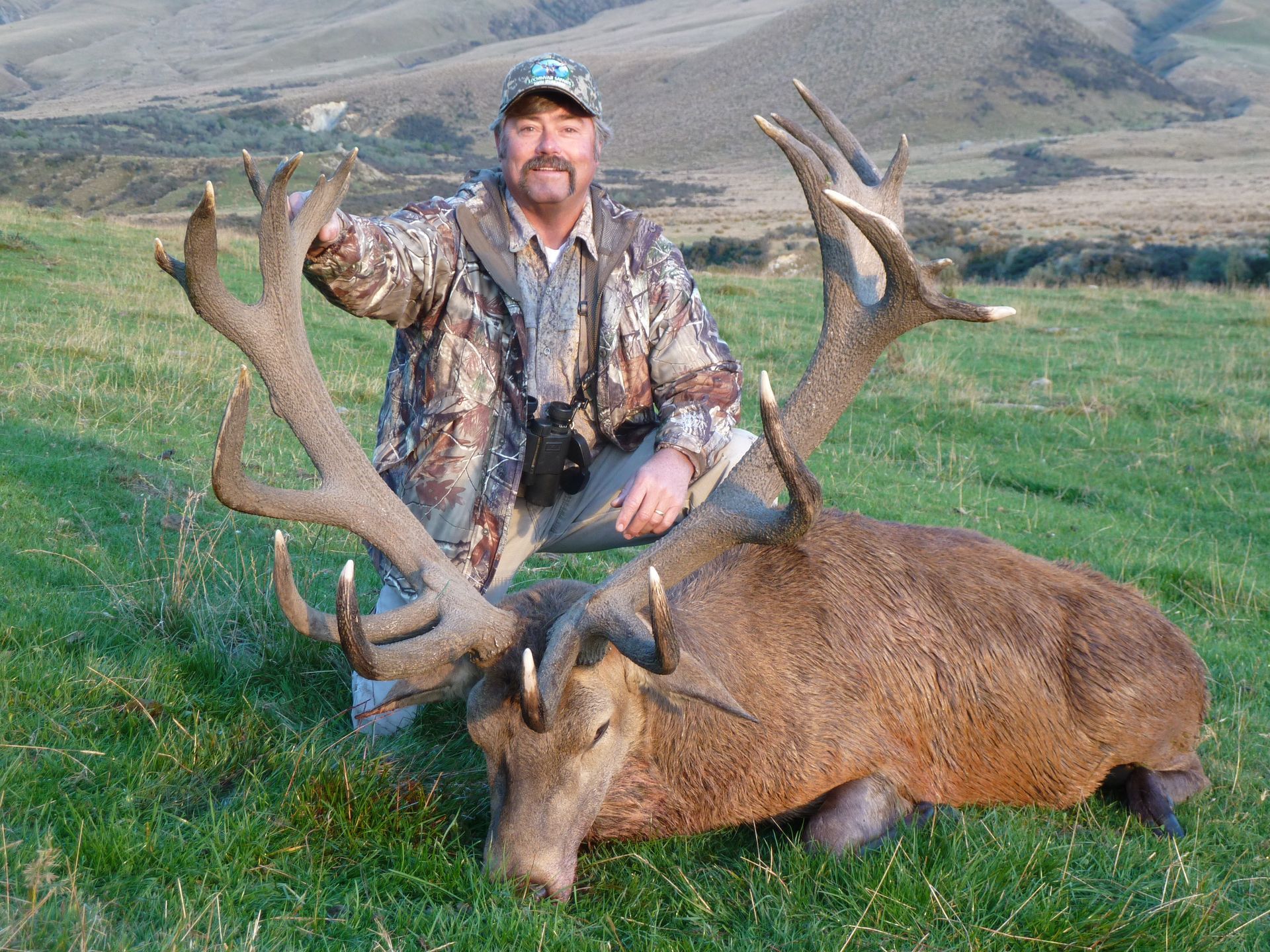 A hunter kneels beside a large dead deer with impressive antlers in a grassy field. The man is wearing camouflage clothing and appears to be smiling.
