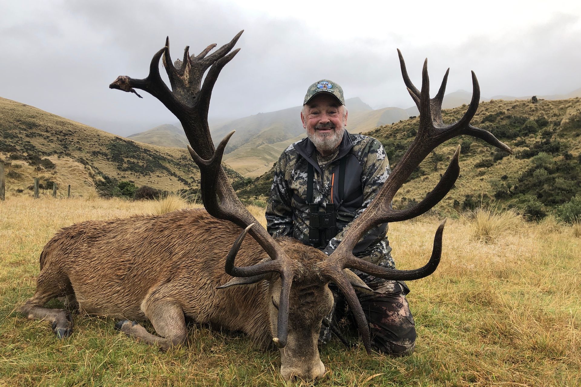 Man kneeling beside a large dead red deer with impressive antlers in a grassy, mountainous landscape. The man is smiling.