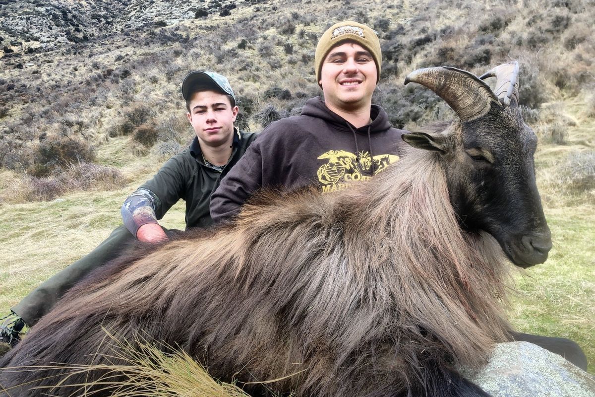 Two men posing with a dead Himalayan tahr in a grassy mountainous area. Both men are smiling, one is wearing a beanie.