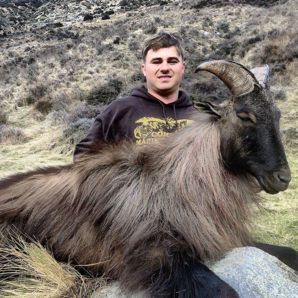 Man in brown hoodie poses with a large, dead Himalayan tahr in a mountainous outdoor setting.