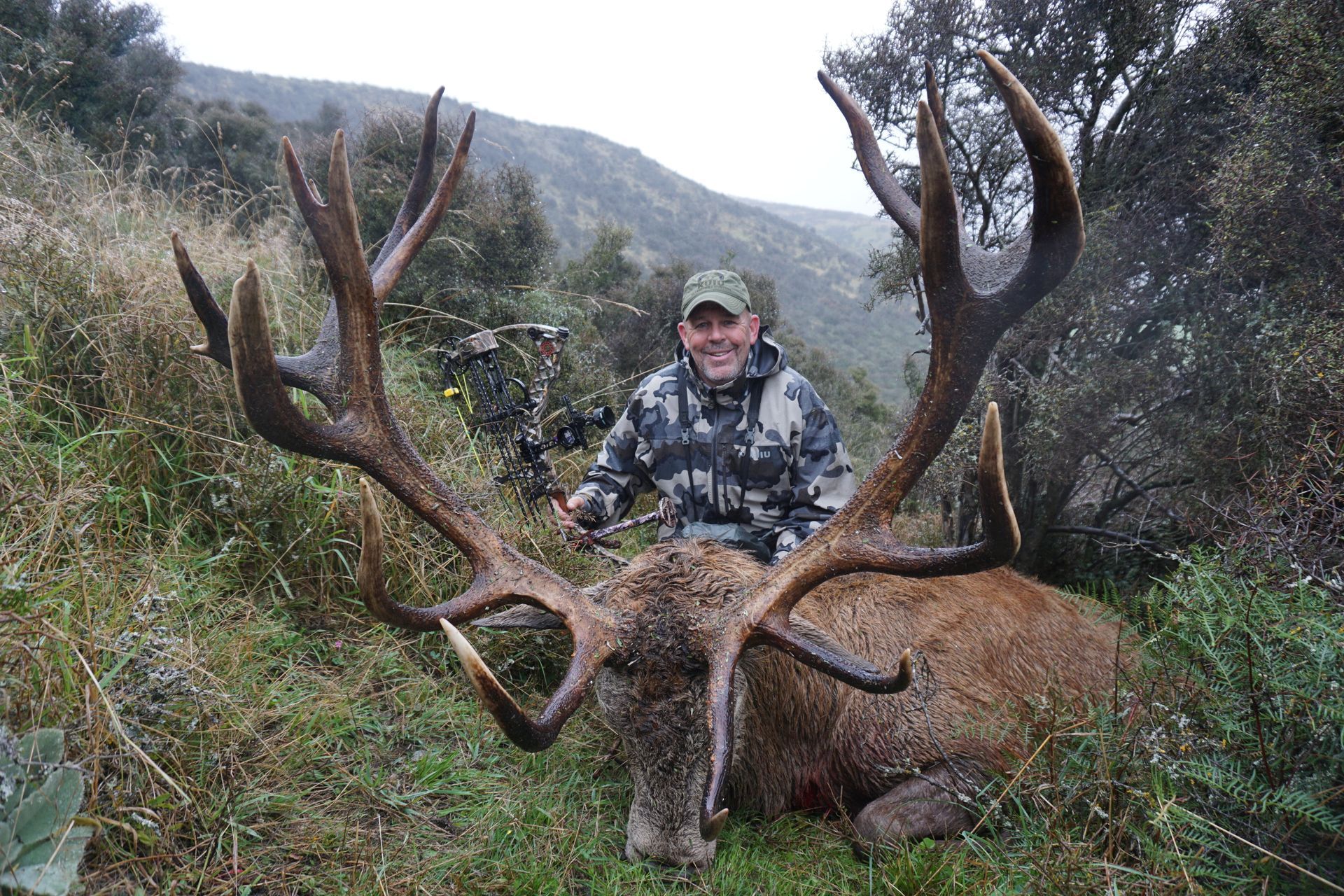 Man in camouflage smiling next to a large red deer with massive antlers, lying in grassy terrain, likely after a hunt.