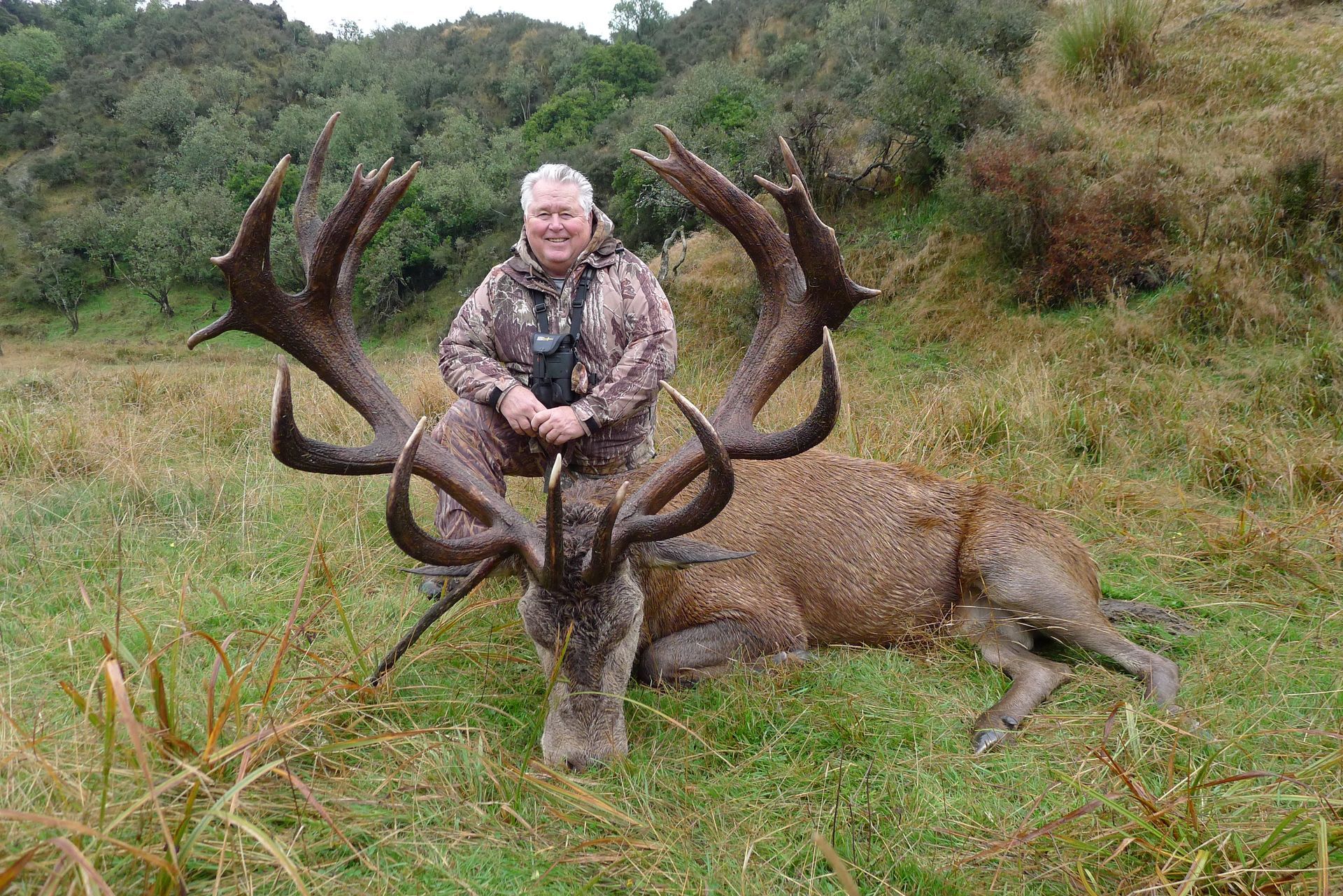Man in camouflage poses with a large dead deer with impressive antlers in a grassy field.