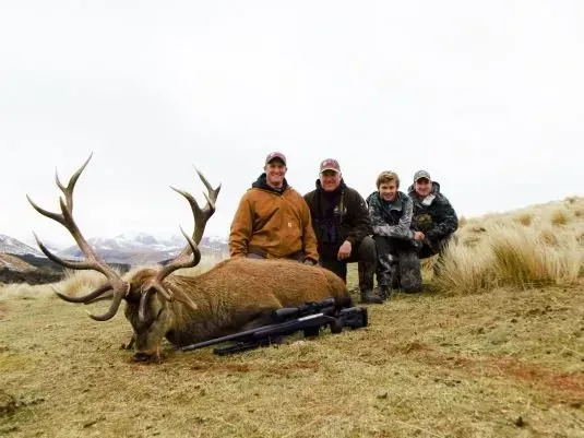 Four hunters pose with a large dead deer with antlers on a grassy hillside. Two rifles lie next to the animal.