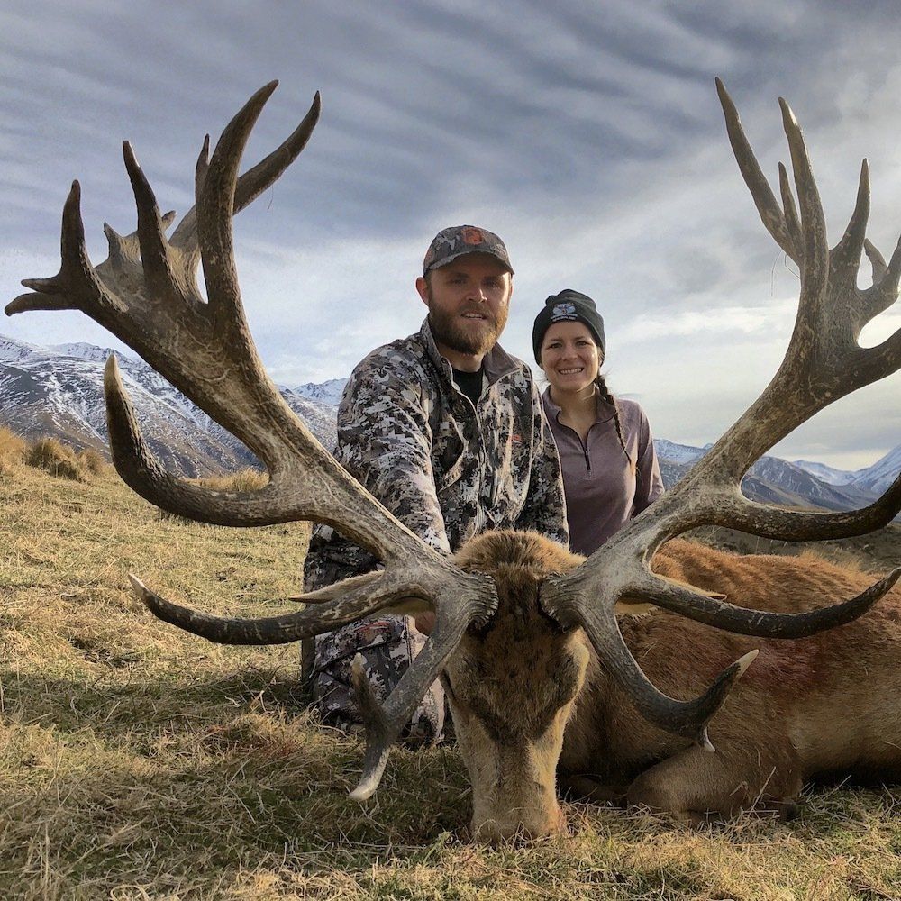 A man and woman kneel next to a large dead deer with impressive antlers in a mountainous outdoor setting.