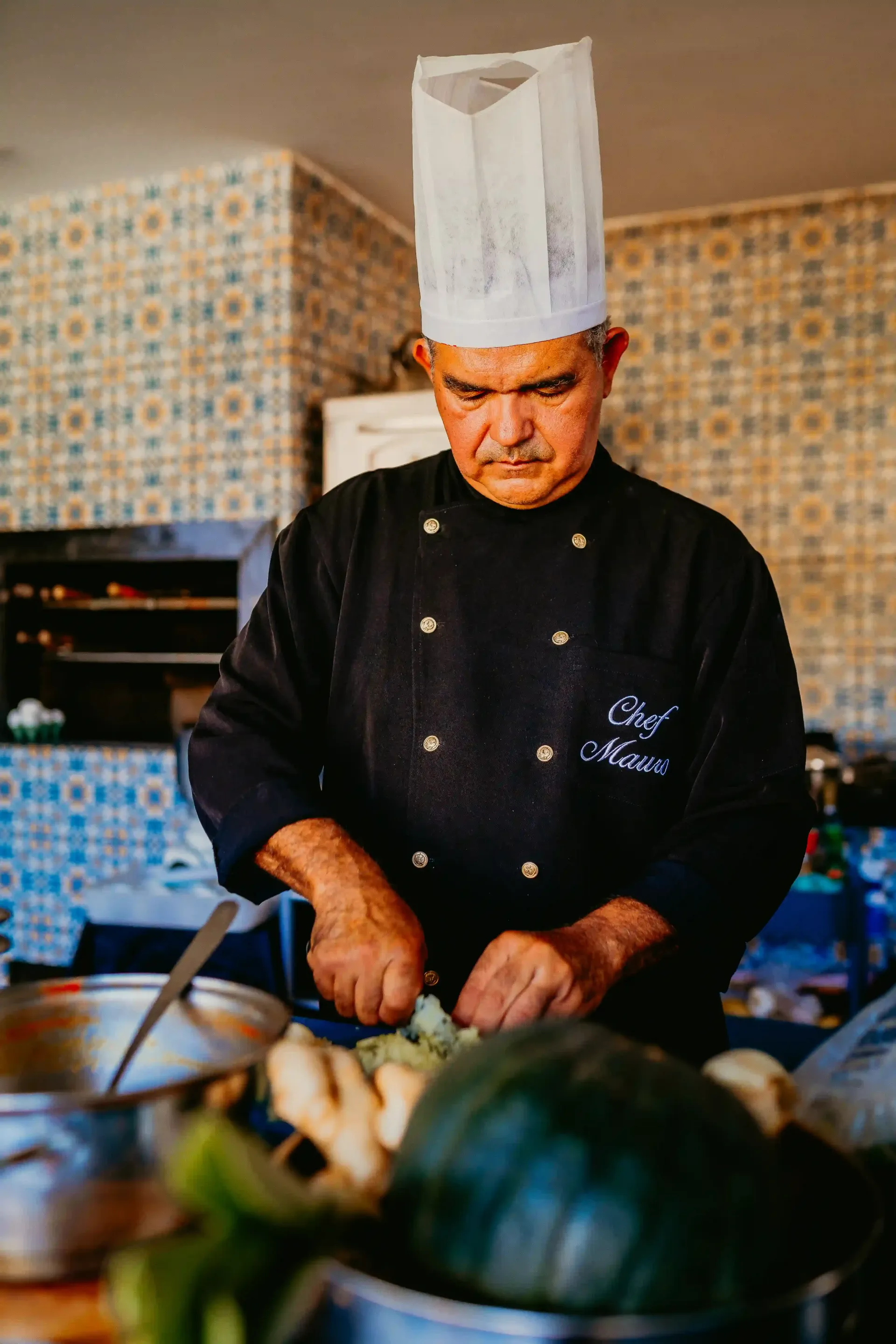 Um homem com chapéu de chef está cortando vegetais na cozinha.-Pousada-Recanto-do-Chef