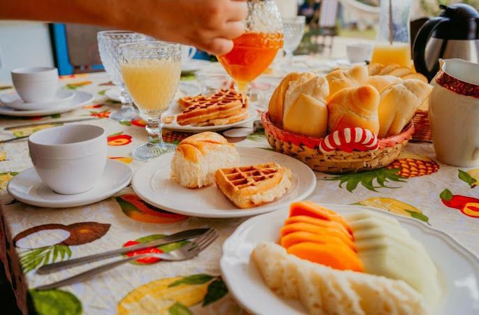 Uma pessoa está despejando suco de laranja em um copo sobre uma mesa com pratos de comida.-Pousada-Recanto-do-Chef