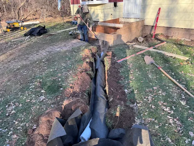 Workers digging a trench beside a house, exposing black drainage pipe and a red-handled shovel.