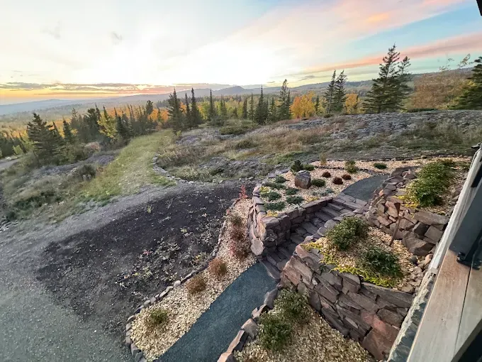 Stone patio and fire pit overlook a grassy hillside with pine trees at sunset.