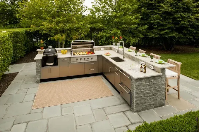 Outdoor stone patio kitchen with grill, sink, counter stools, and potted plants in a garden setting