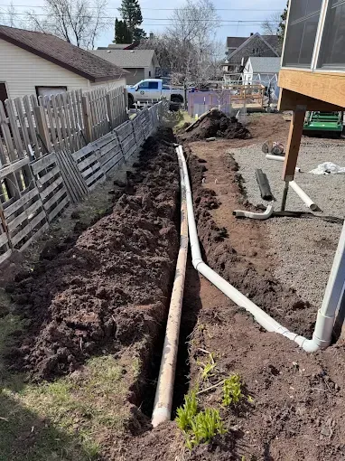 Trenched backyard with exposed pipes and dirt piles beside a fence and house under construction