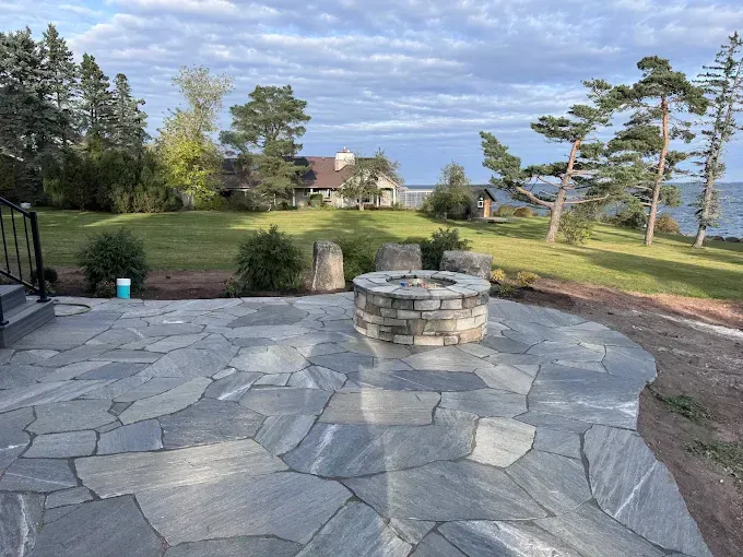 Stone patio with round fire pit overlooking a grassy yard and water under a cloudy sky
