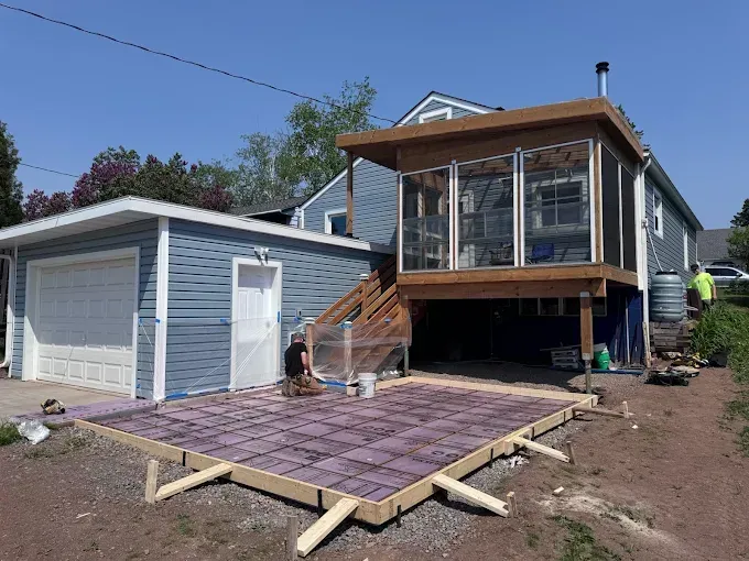Backyard deck under construction beside a house, with a worker and framed patio foundation.