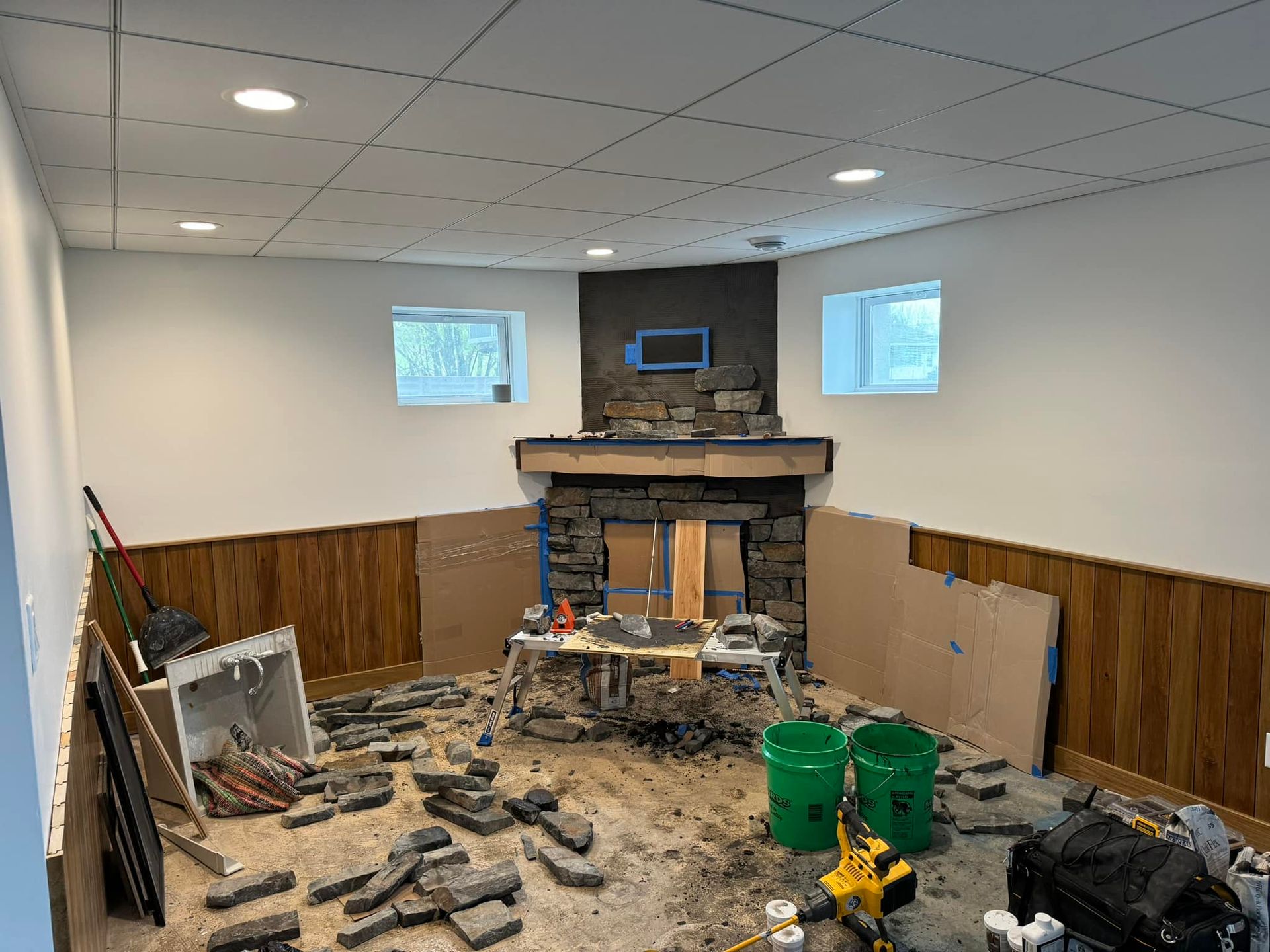 Partially renovated room with debris on the floor, stone fireplace, and wood paneling on the walls