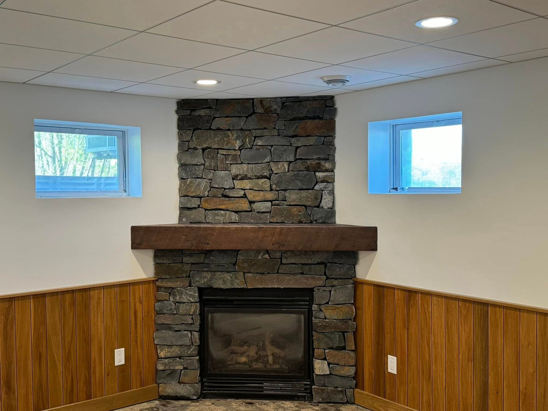Basement room with stone fireplace, wood mantel, and two small windows above wood-paneled walls.