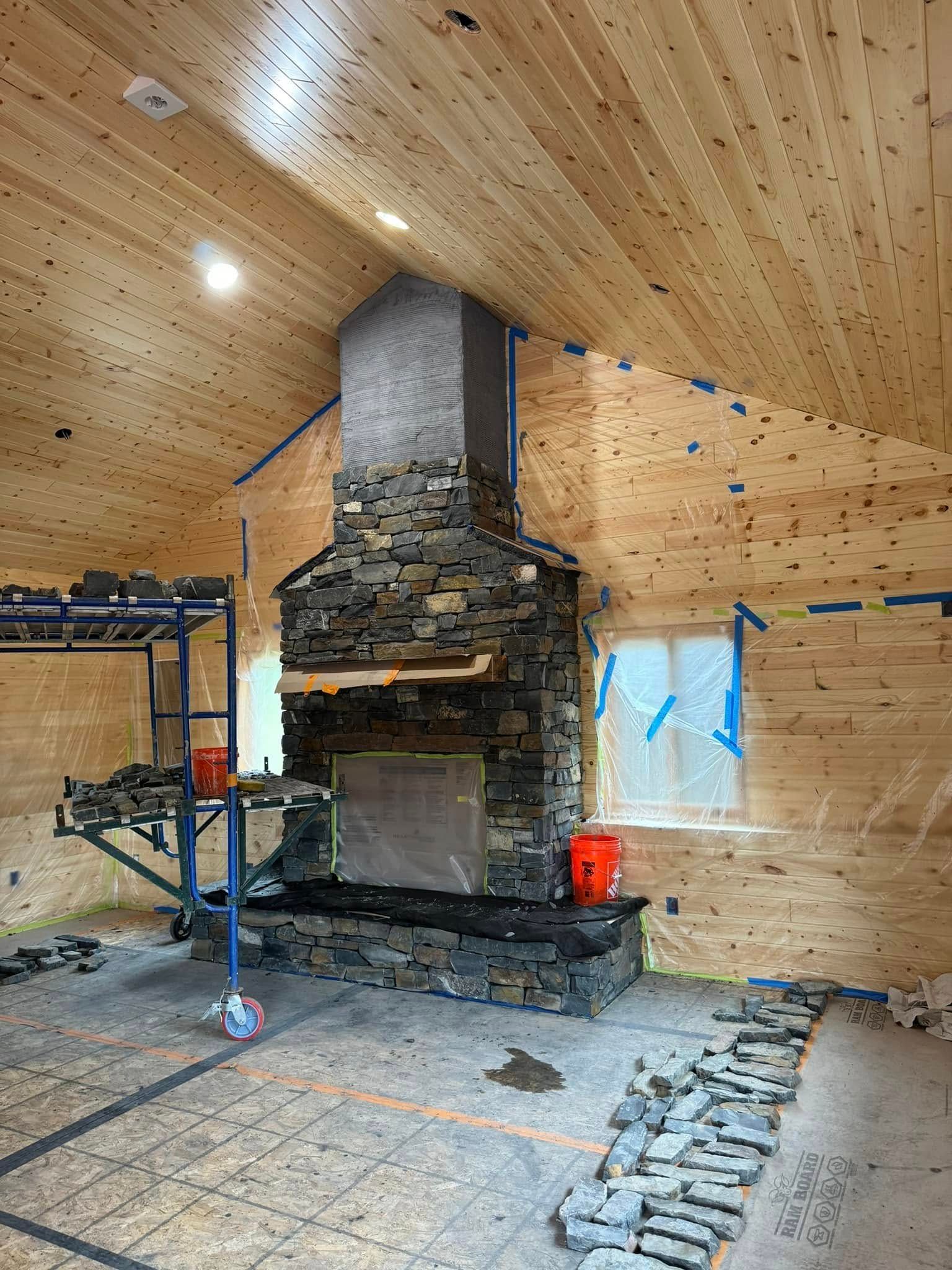 Rustic room with wood-paneled ceiling and walls, central stone fireplace, and plastic-covered windows under construction
