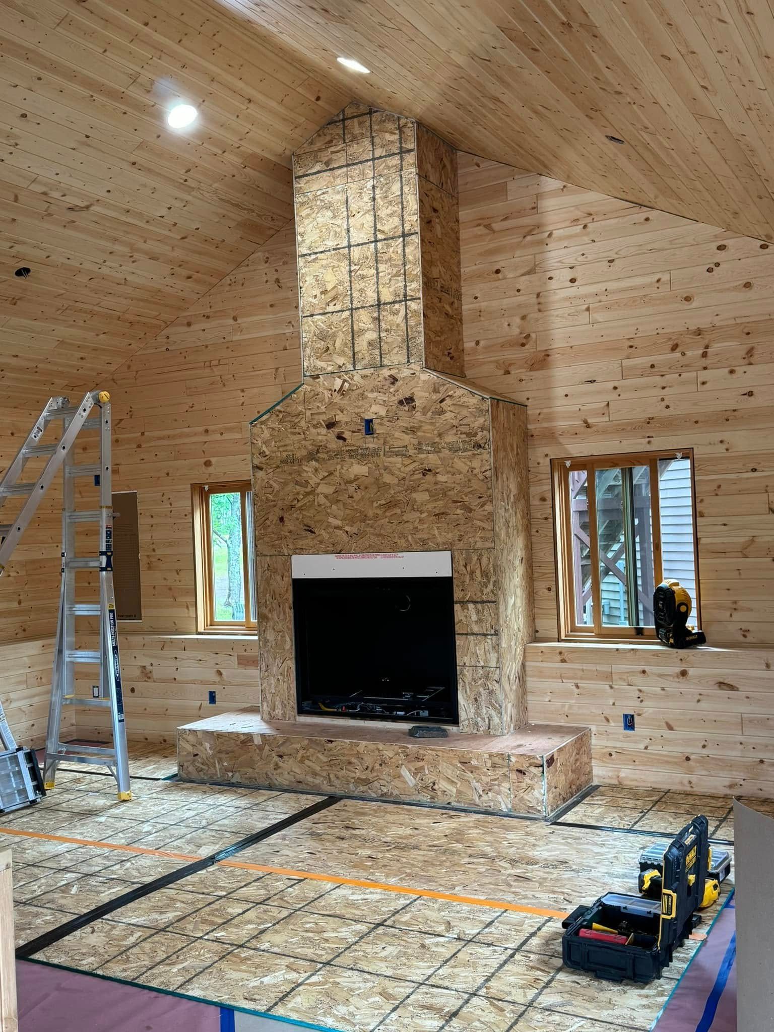 Unfinished wood-paneled room with a stone fireplace, ladder, and tools on the floor.