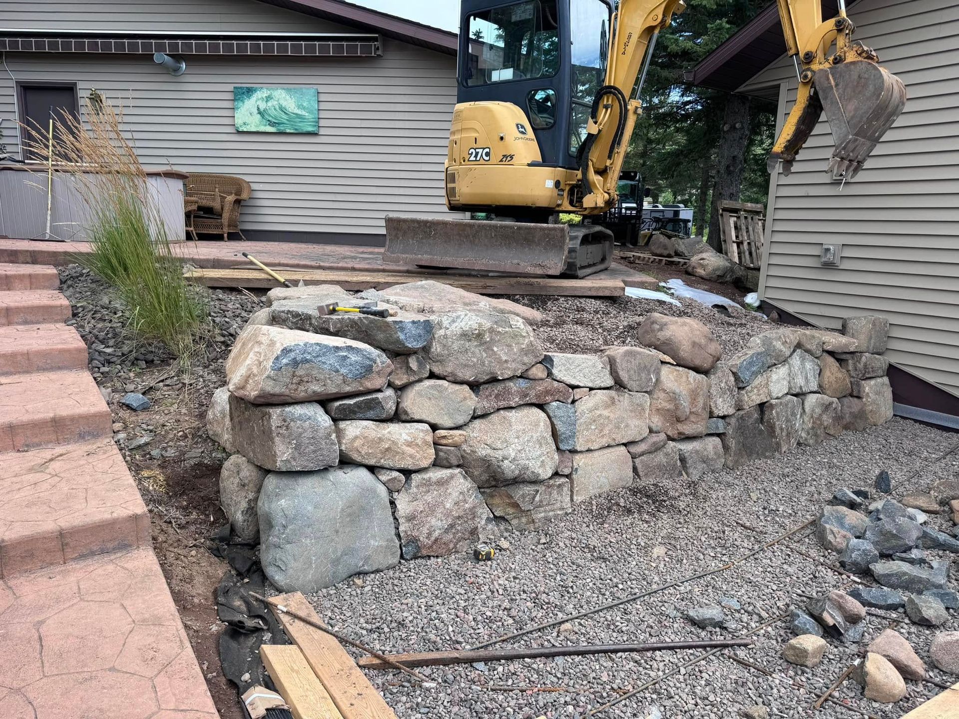 Excavator works beside a stone retaining wall under construction near a house and walkway.