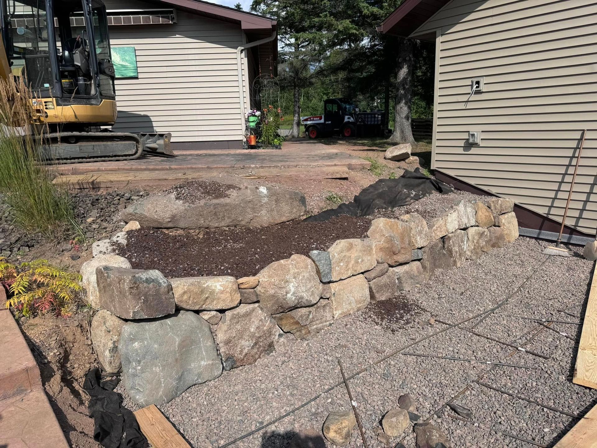 Stone retaining wall and gravel patio under construction between two beige houses, with an excavator nearby