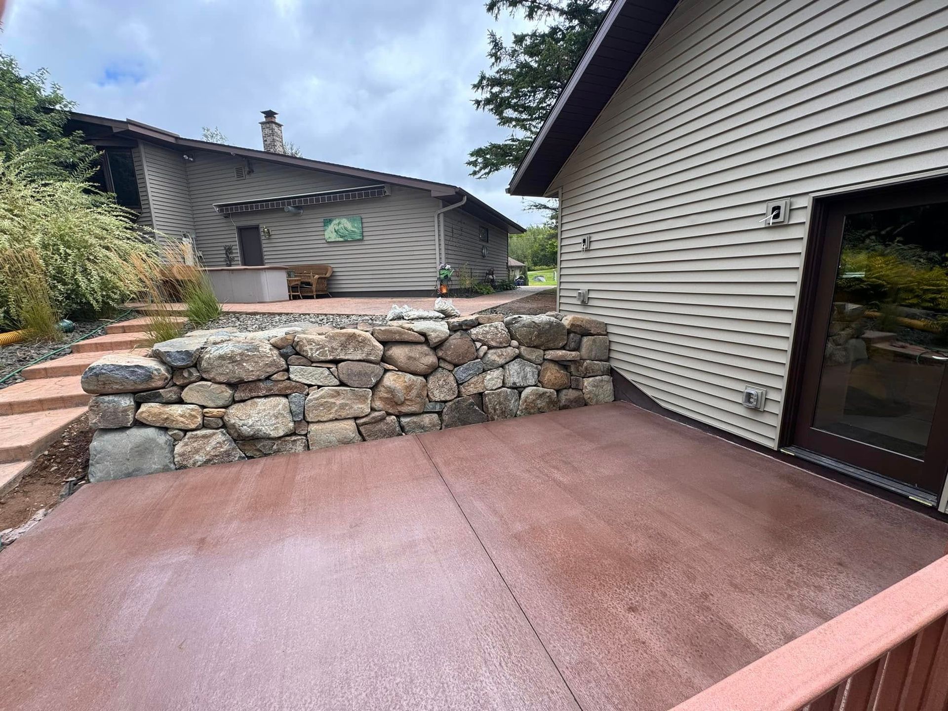 Stone retaining wall beside a red concrete patio and beige house exterior with a covered opening