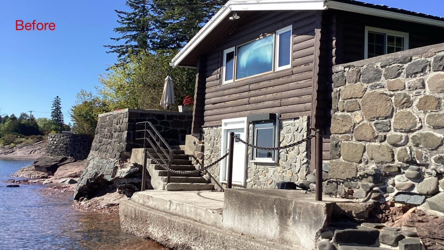 Lakefront stone and wood house with stairs and seawall under a clear blue sky.