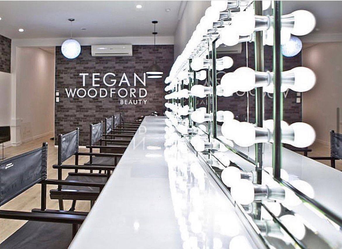 A row of chairs in a salon with the words tegan woodford beauty on the wall