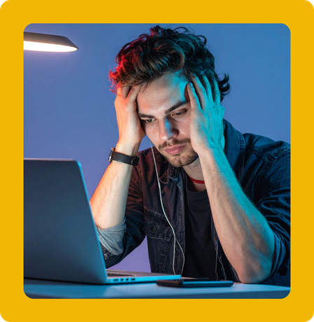 Man stressed at laptop, hands on head, with earphones, illuminated by desk lamp, blue and red lighting.