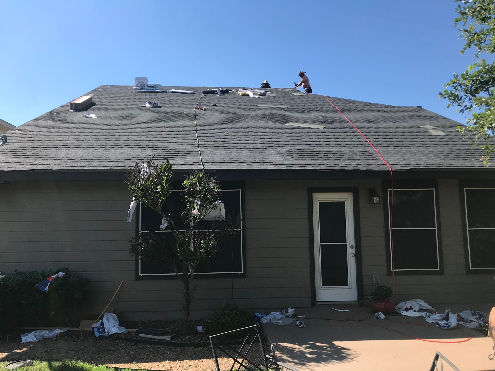 Roofers working on a dark shingled roof under a blue sky. House with green siding and open door.