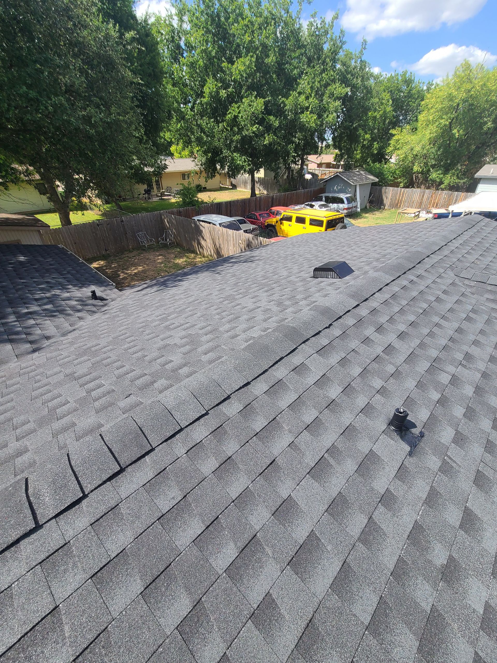 View of a gray shingle roof with a yard and trees in the background.