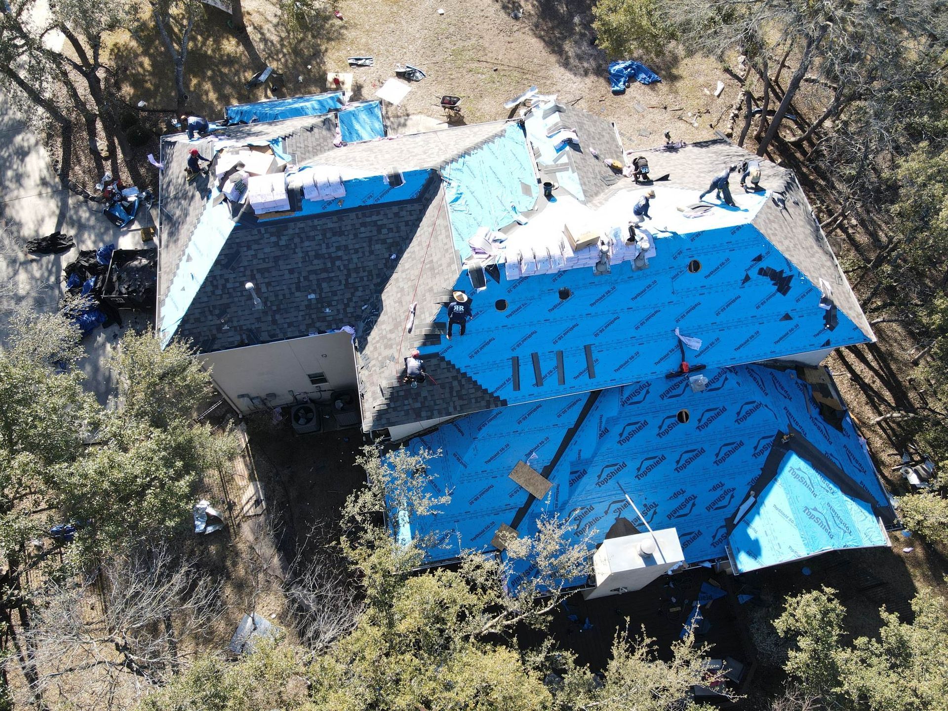 Aerial view of a house undergoing roofing work, showing blue underlayment and workers on the roof.