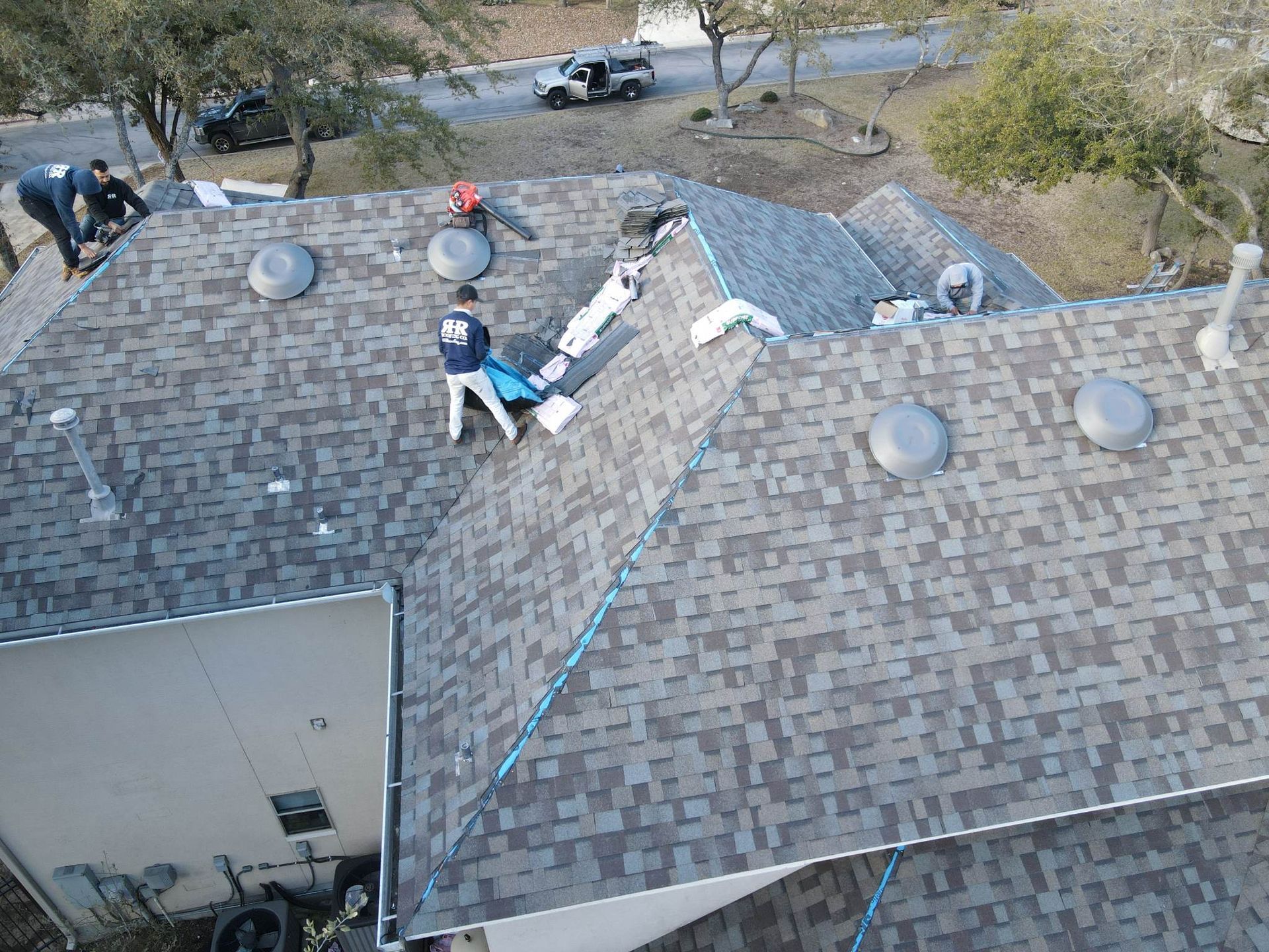 Workers on a roof with grey shingles, wearing safety gear. Outdoors, sunny day.
