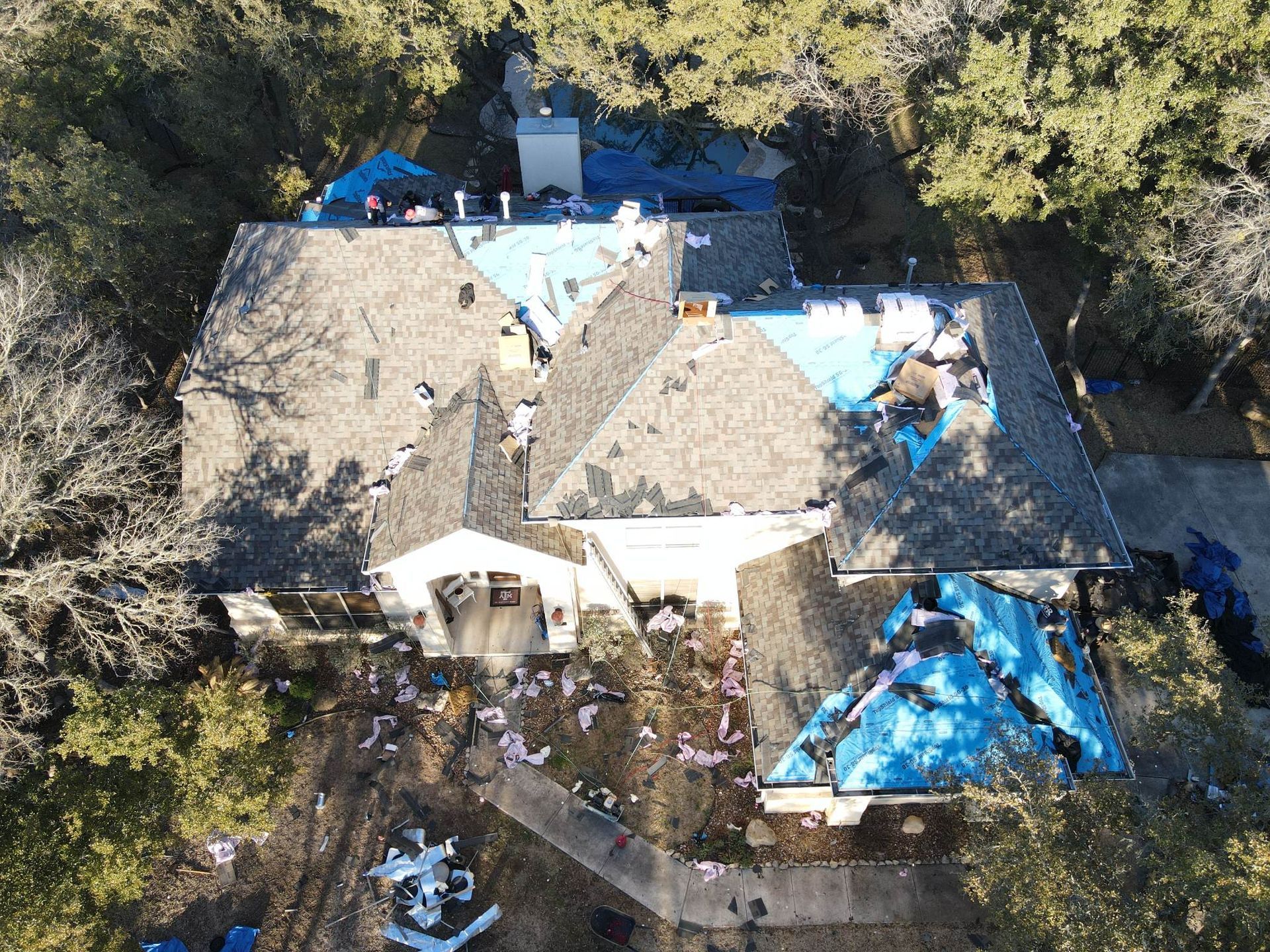 Aerial view of a house with a damaged roof, covered with blue tarps. Debris scattered on the ground.