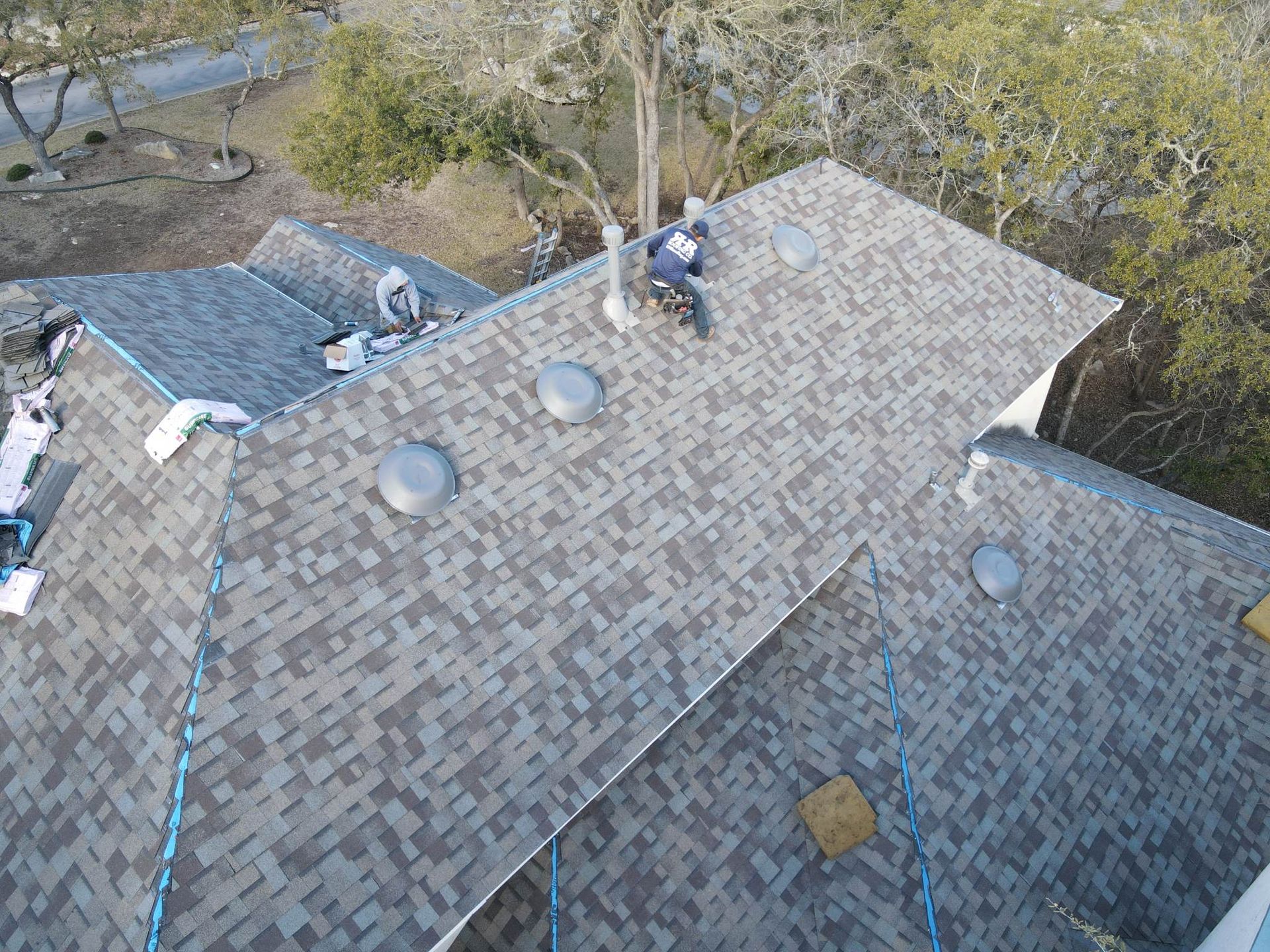 Roofers installing shingles on a residential roof. Workers are near a chimney and vents.
