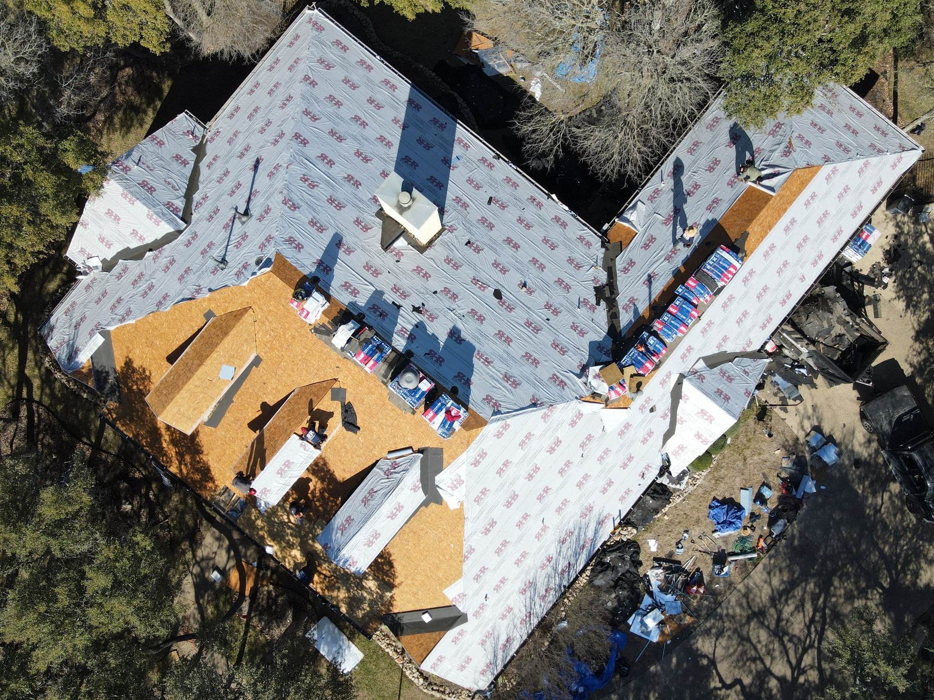 Aerial view of a house with a partially replaced roof; workers are installing shingles, with materials scattered around.