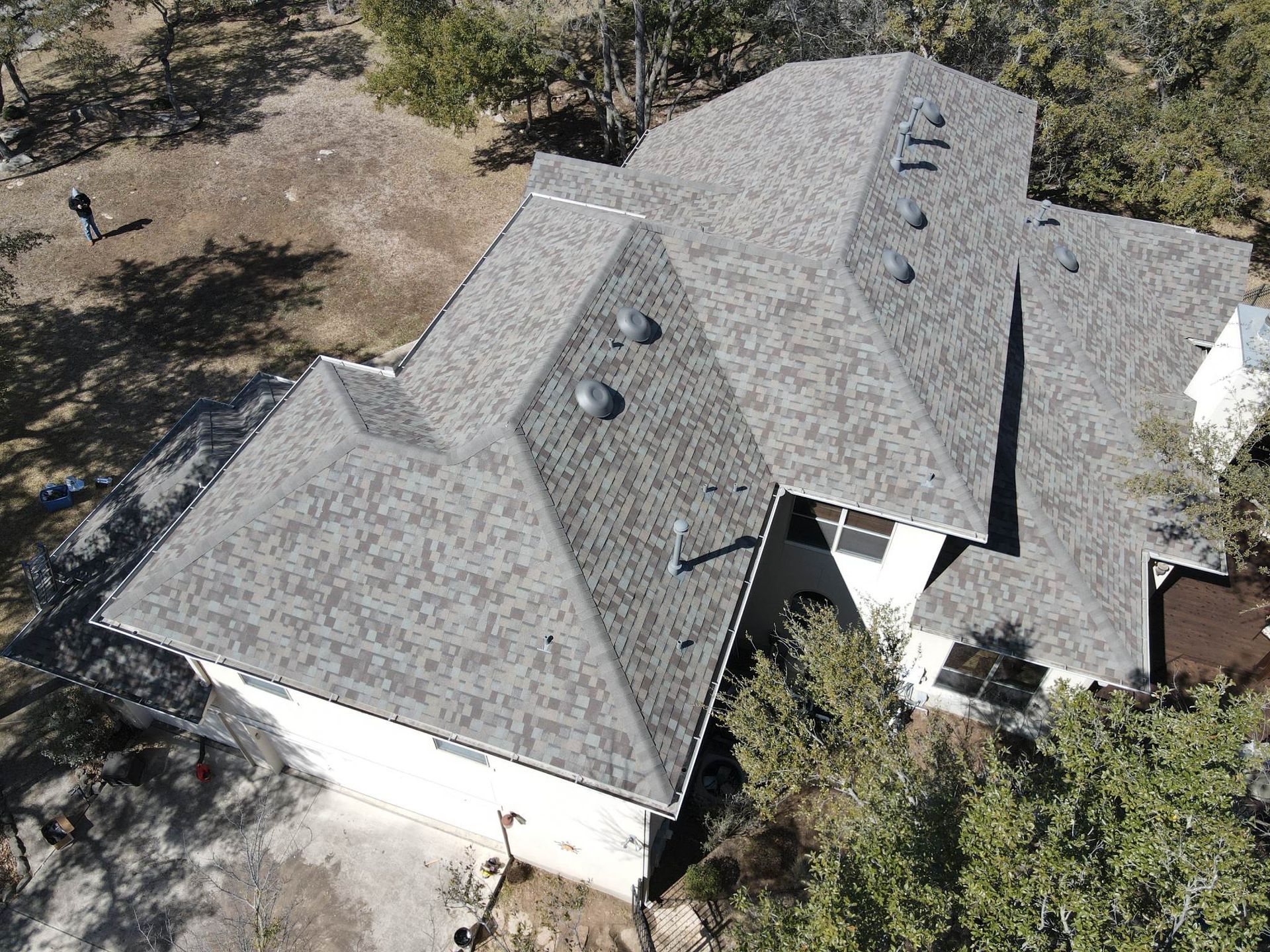 Aerial view of a house with a gray roof surrounded by trees and a person standing nearby.