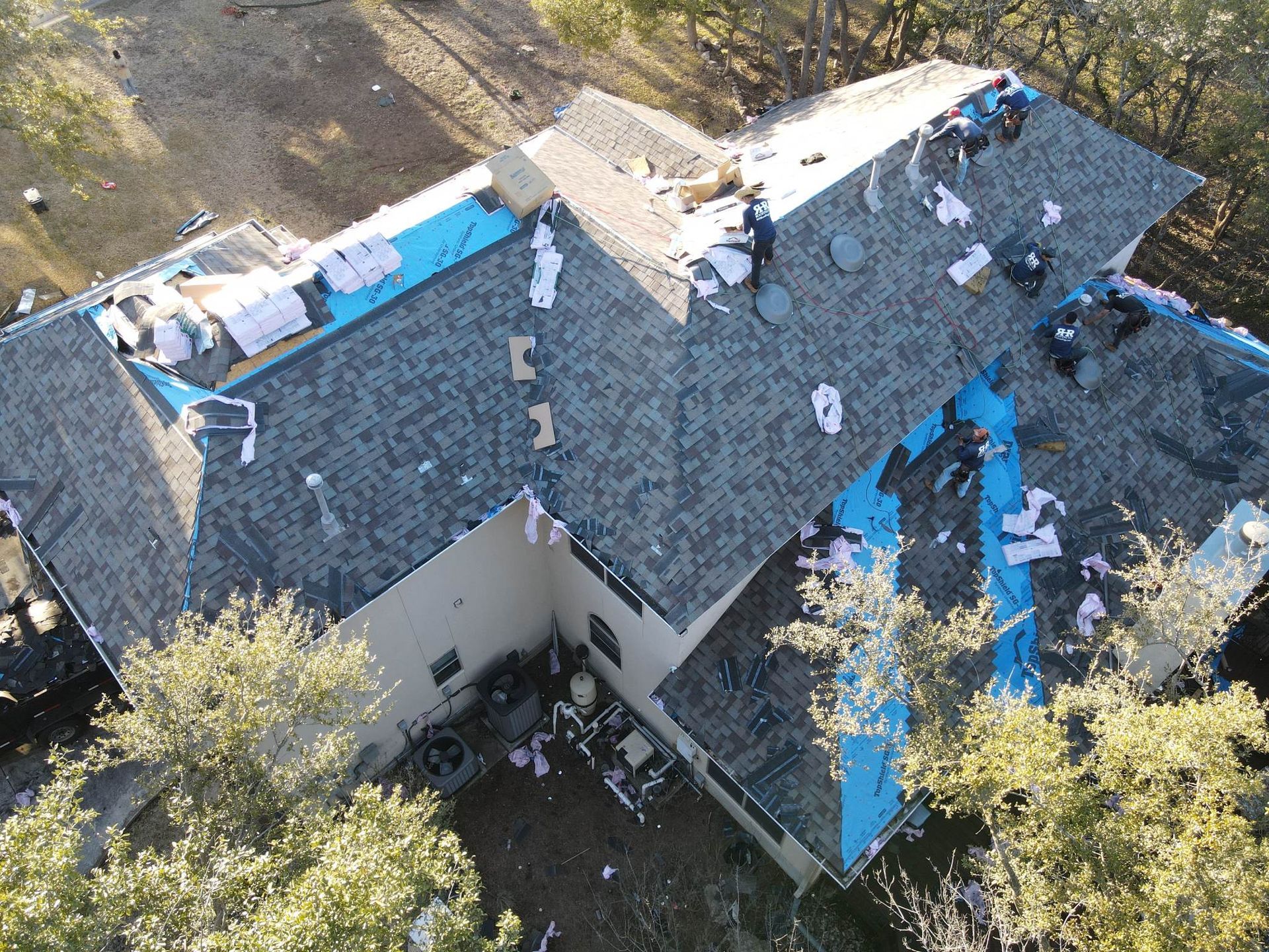 Aerial view of a house roof in disrepair with blue tarp covering exposed areas and workers.