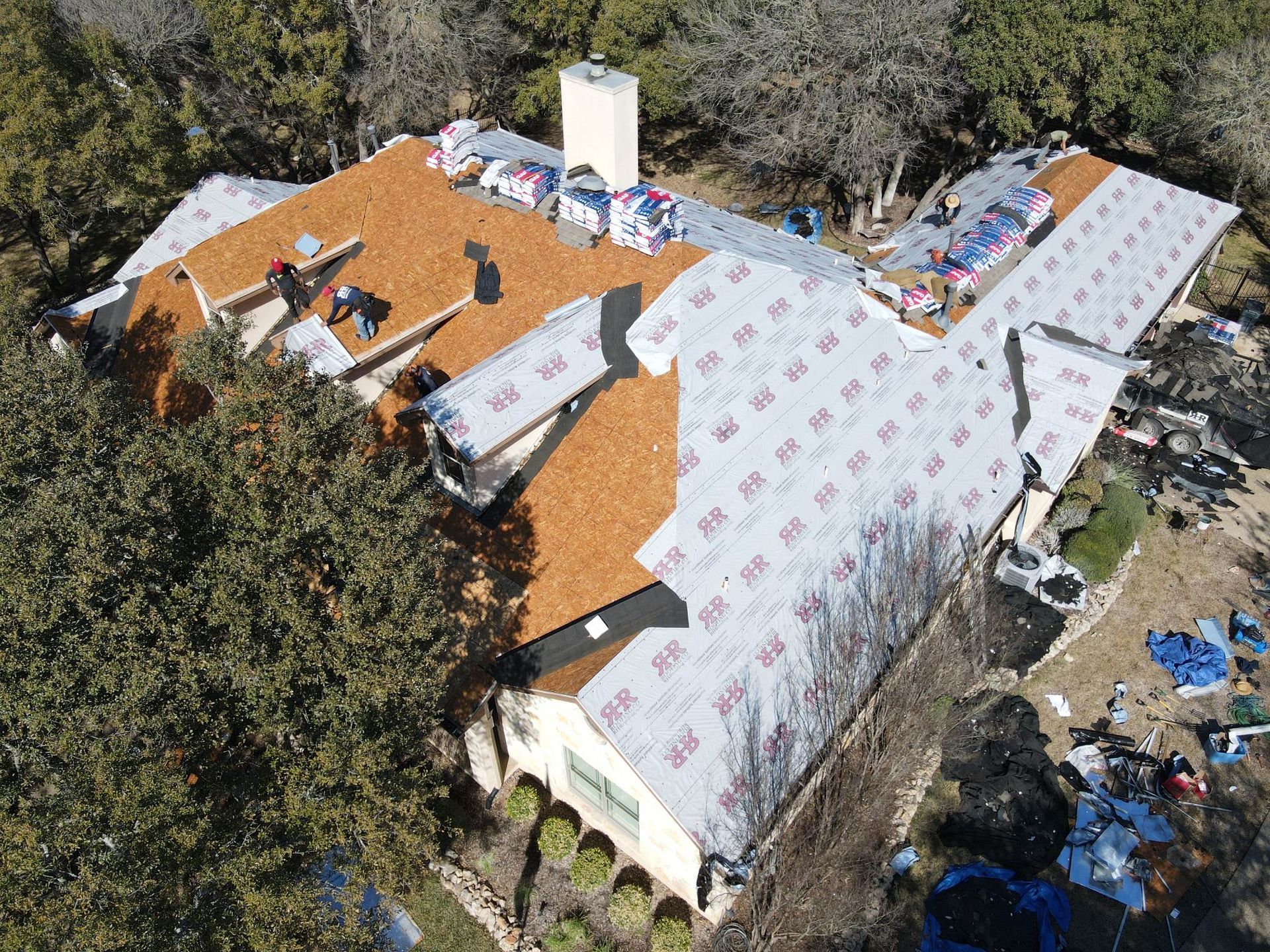 Aerial view of a house roof under construction, workers applying brown shingles over gray underlayment.