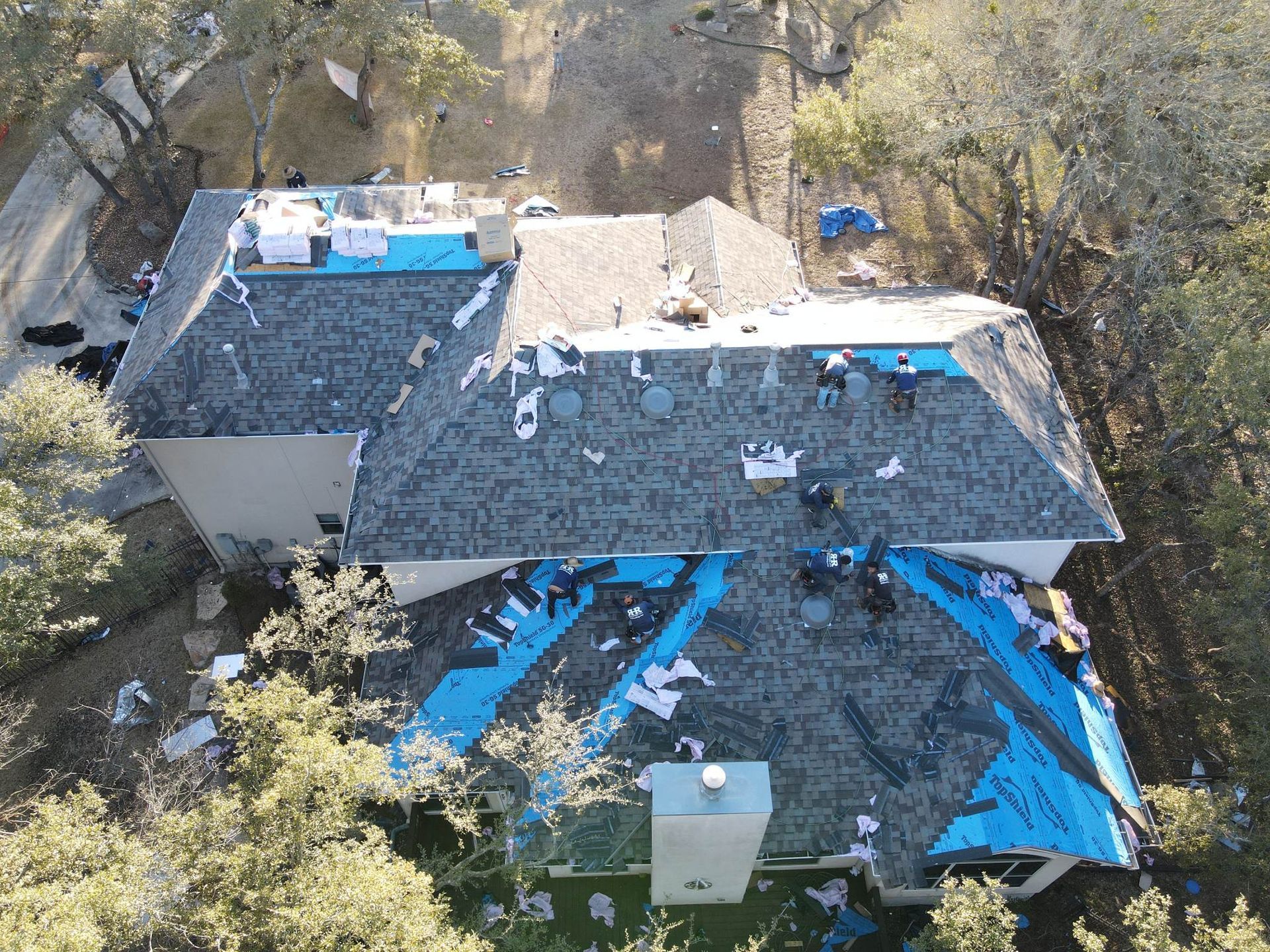 Roofers working on a house with blue underlayment visible. Debris and trees surround the home.