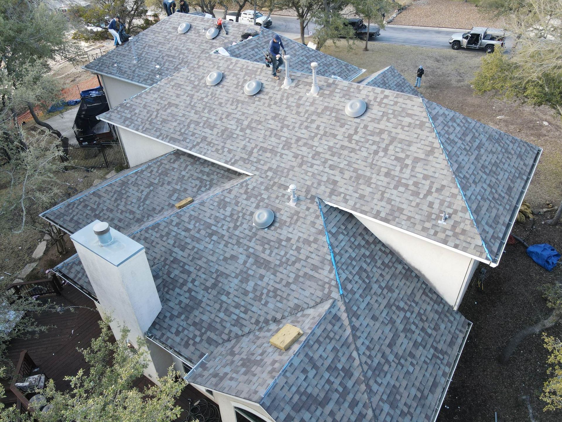 Workers installing a new gray shingle roof on a residential house with several roof vents and a white chimney.