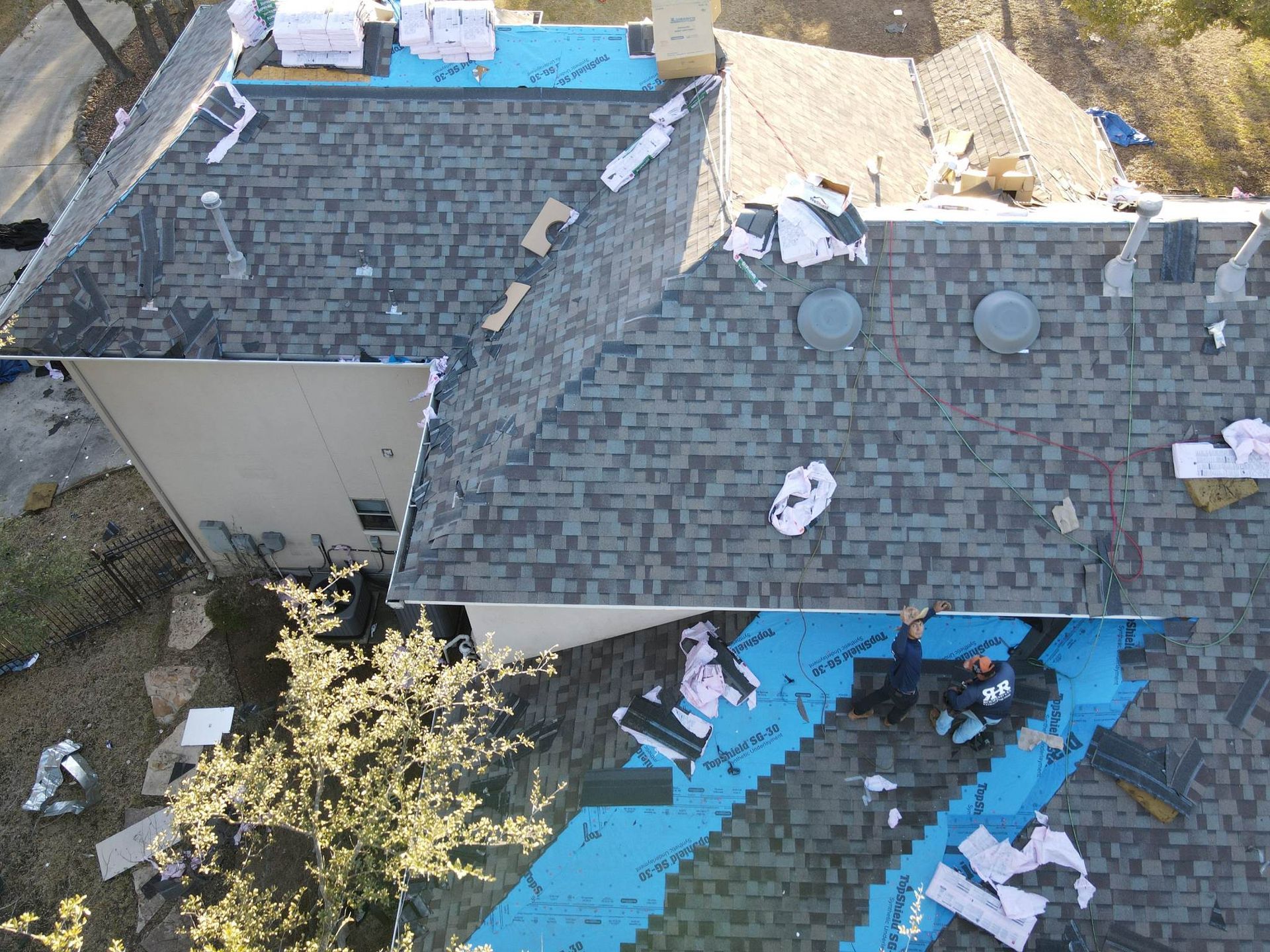 Aerial view of a house under roof repair. Workers on the roof, shingles and blue underlayment visible.
