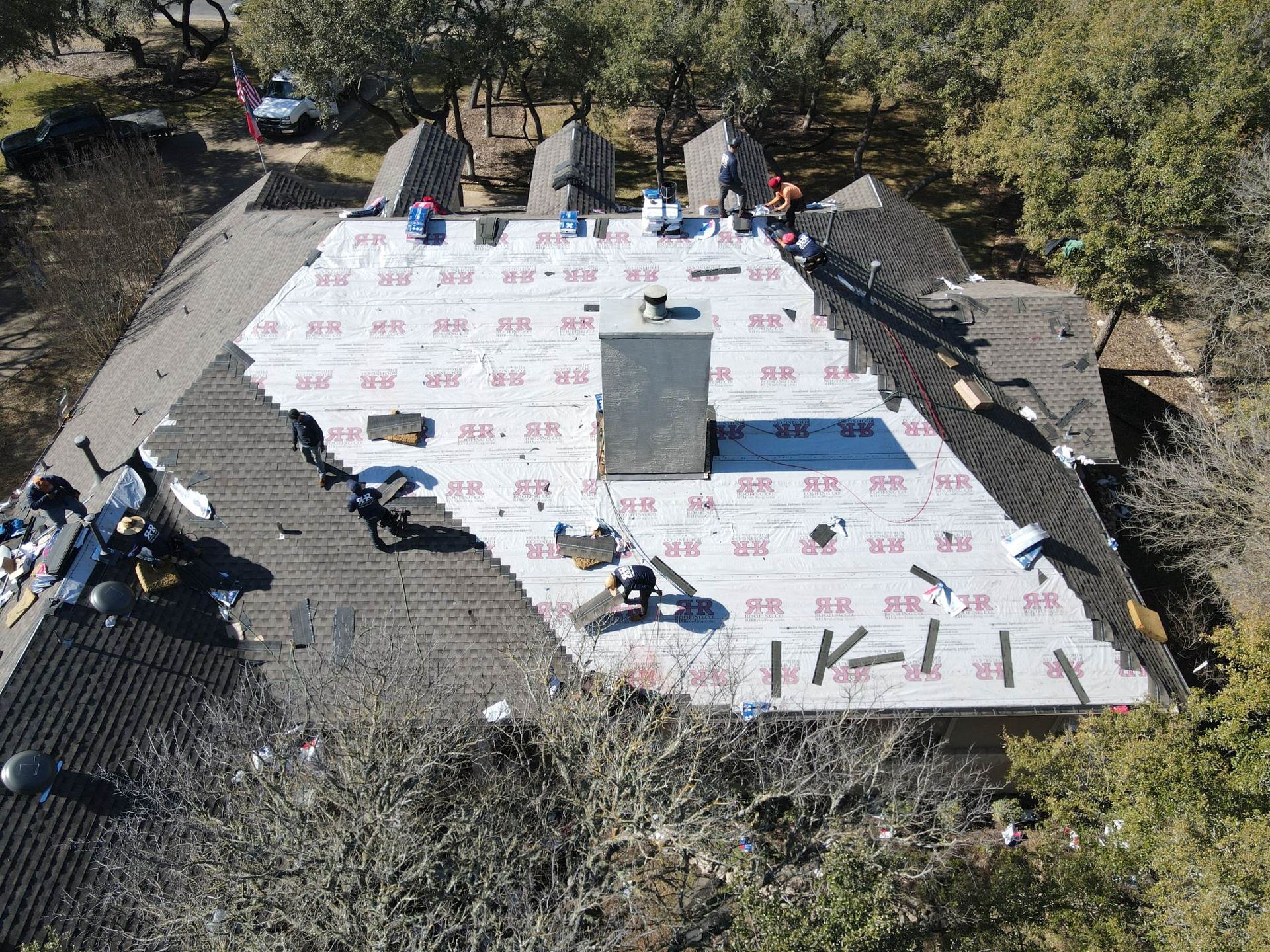 Roofers replacing shingles on a house with a chimney and surrounding trees.