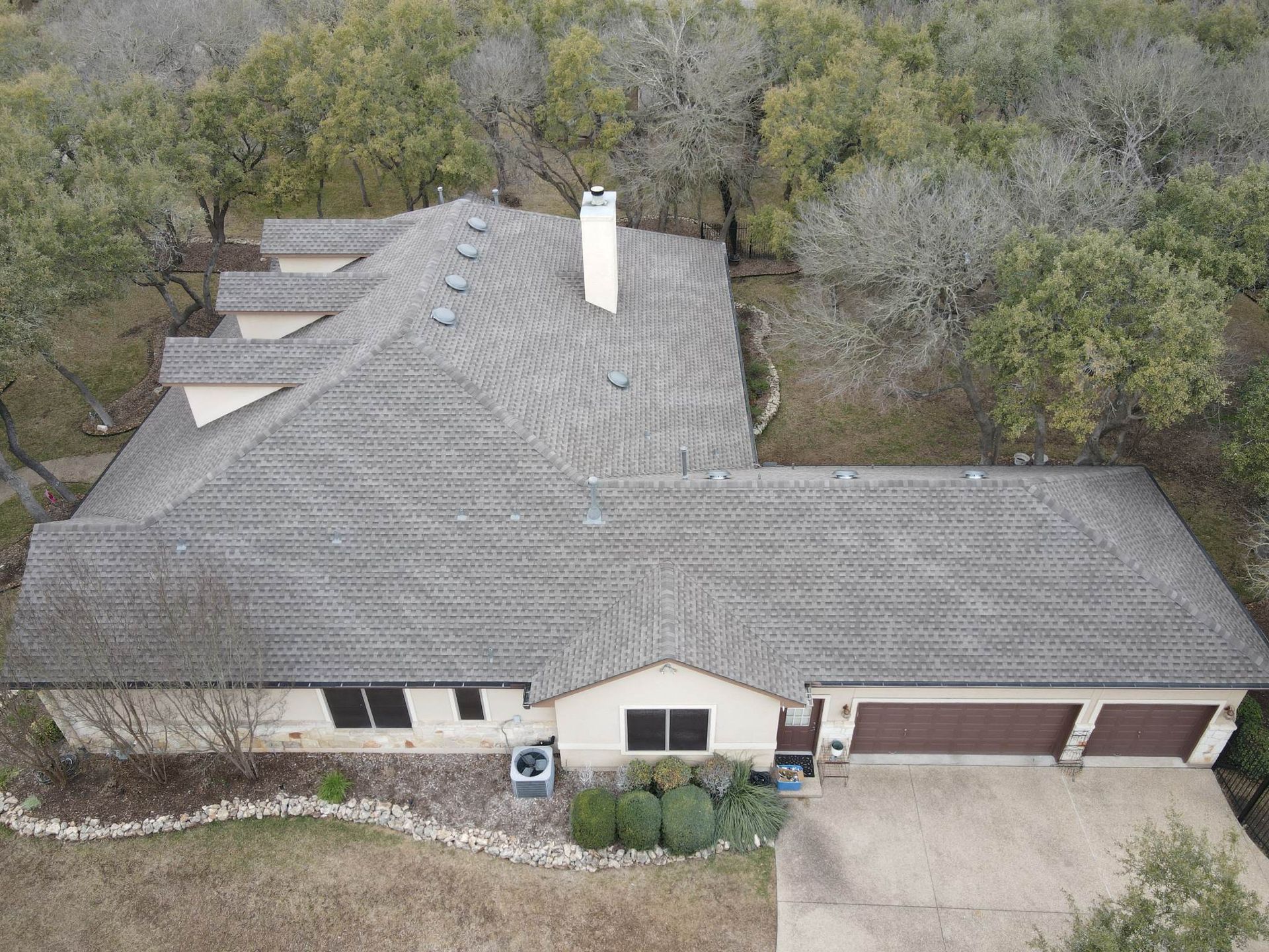 Aerial view of a house with a gray roof, beige walls, and brown garage doors surrounded by trees.