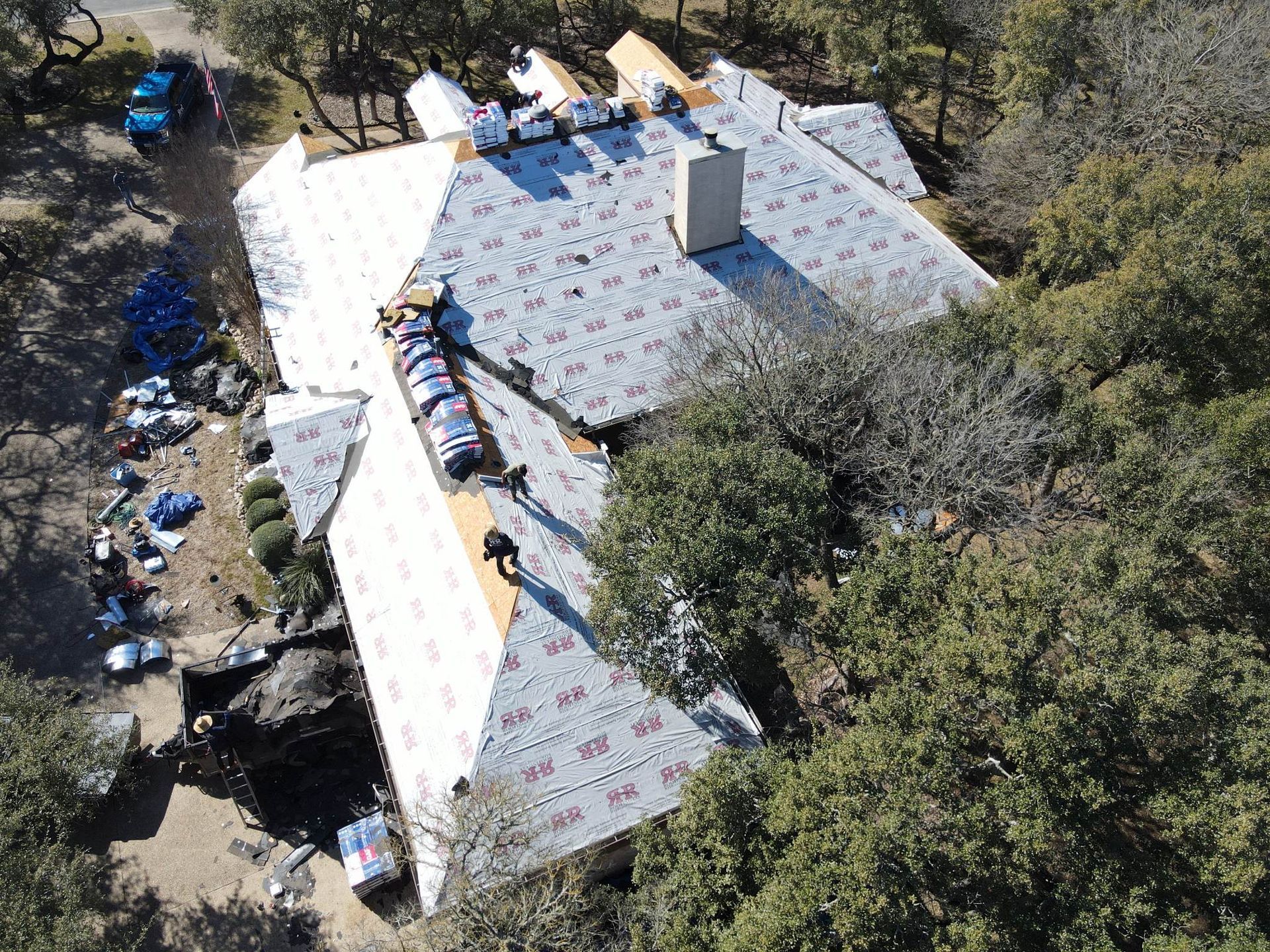 Aerial view of a house being re-roofed; workers, rolls of roofing material, trees surround the house.