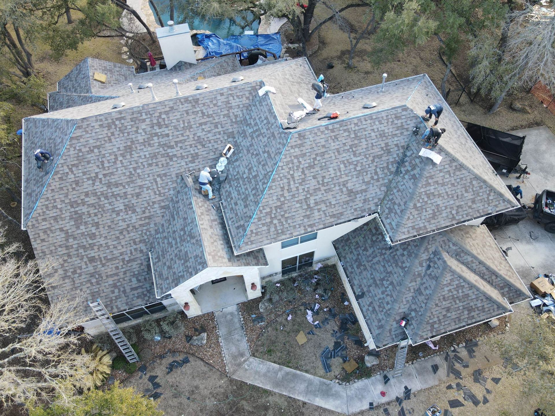 Roofers working on a gray shingle roof, surrounded by debris. House with white walls, walkway. Aerial view.