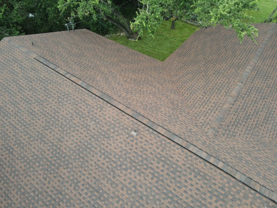 Brown asphalt shingle roof with a center channel. Green grass and trees in the background.