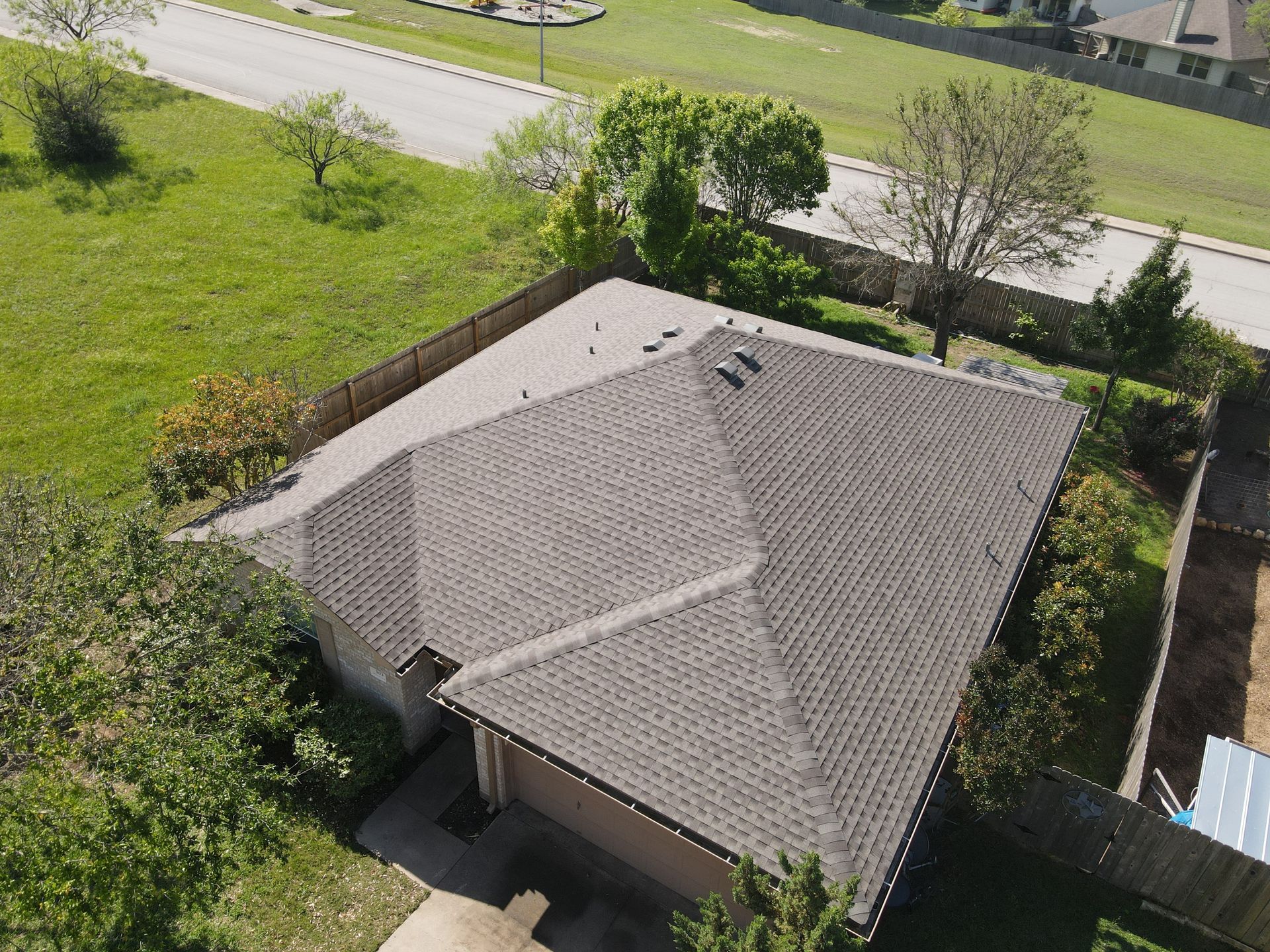 Overhead view of a house with a brown roof, surrounded by green grass and trees; a street is visible in the background.